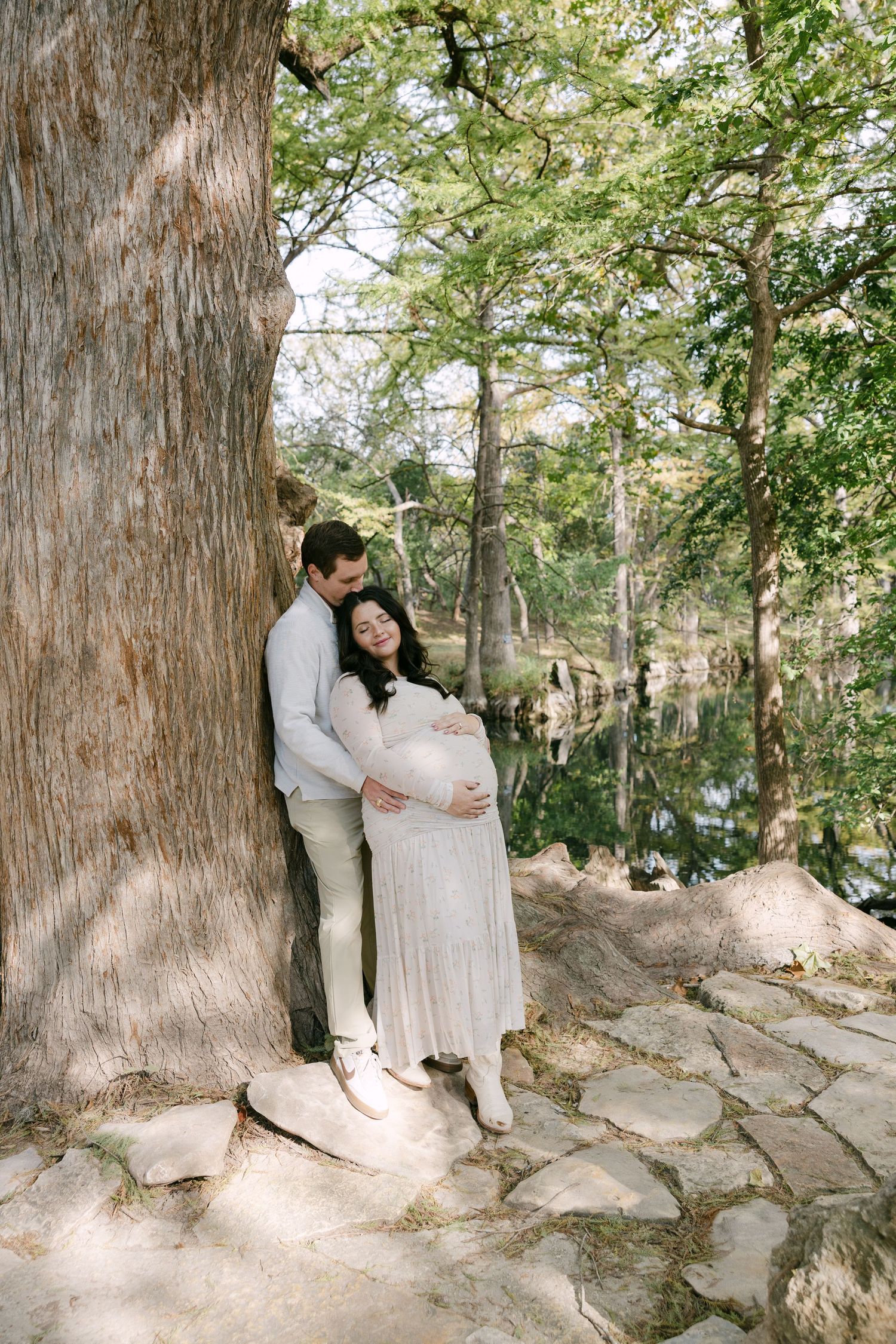 Couple embracing by a large tree near a reflective pond during maternity photoshoot.