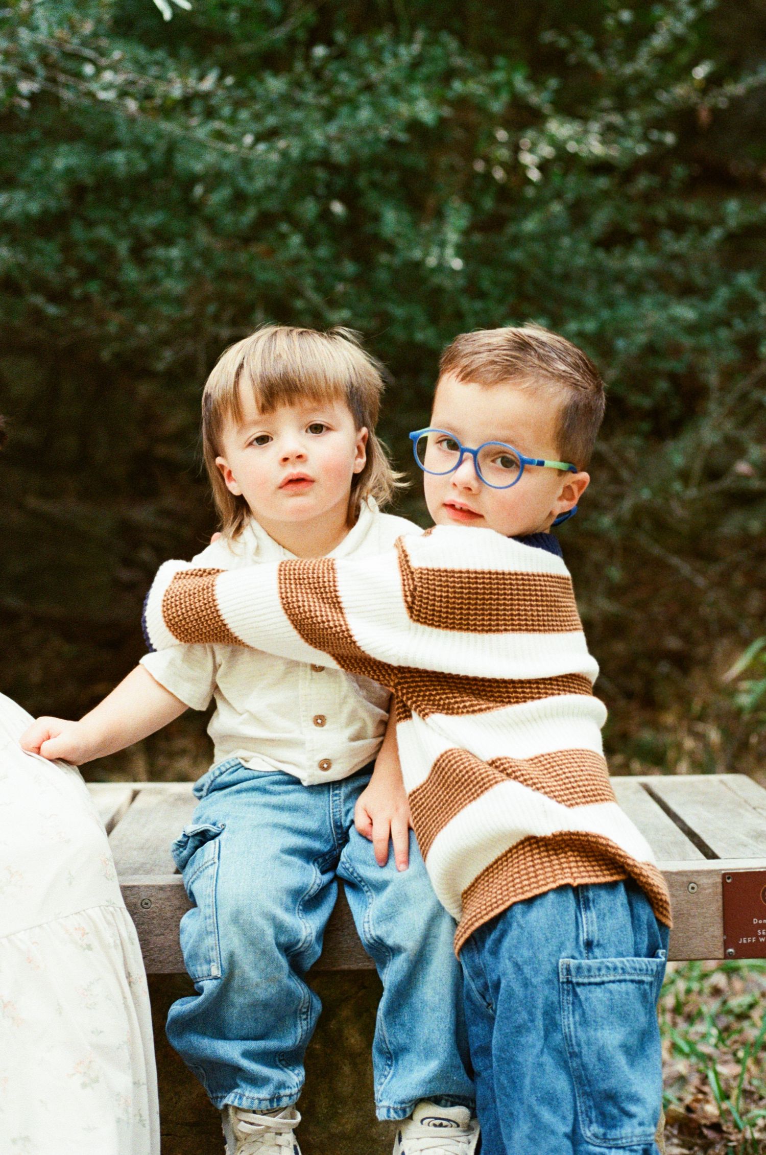 Two young boys in matching striped sweaters sitting together on a wooden bench outdoors.