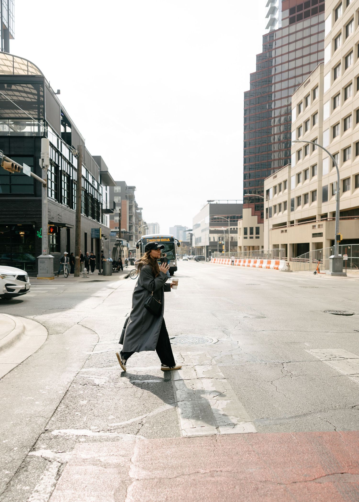 Woman in long black coat crossing urban street while holding camera in city.