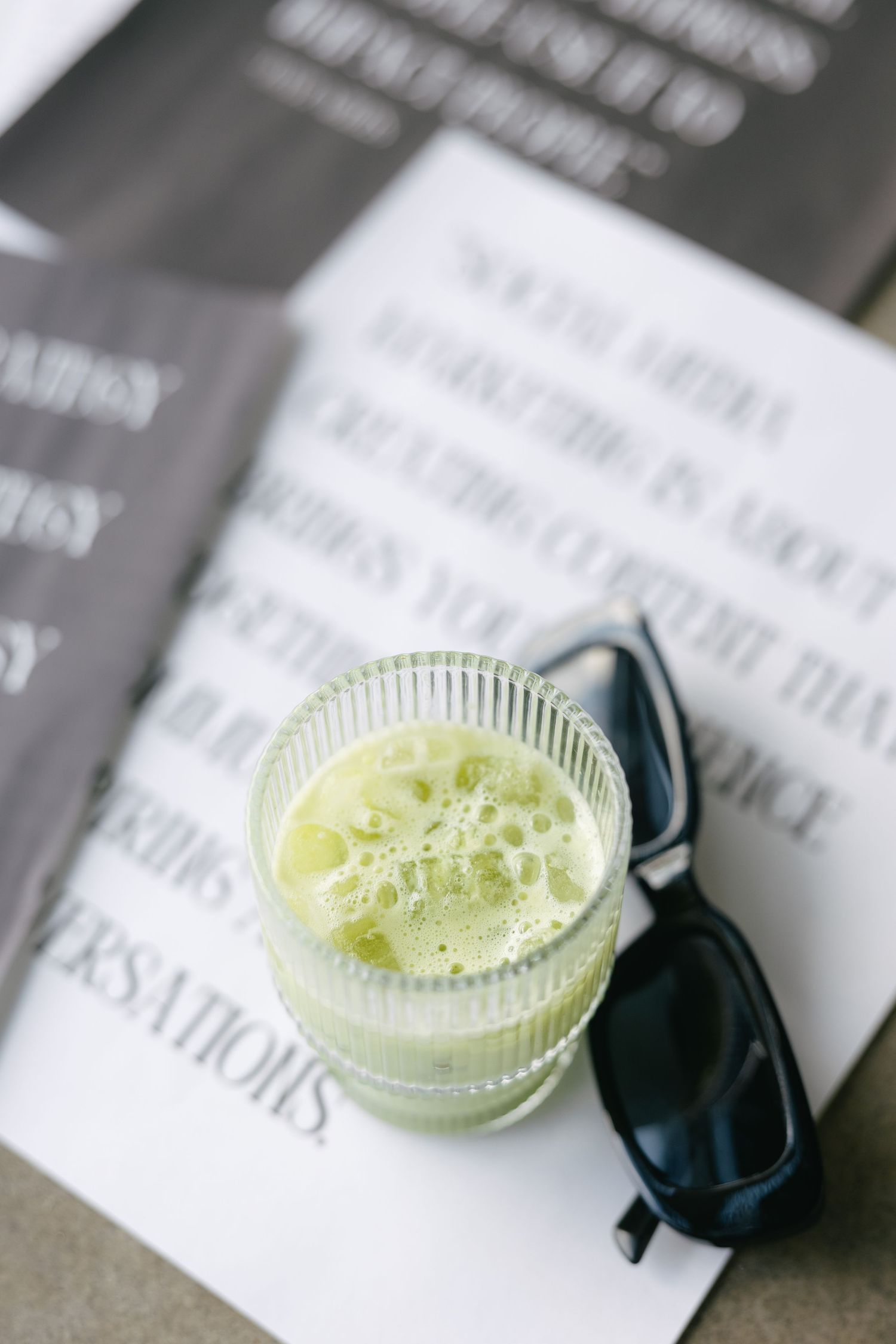 Green matcha drink in ribbed glass beside black sunglasses on printed paper.
