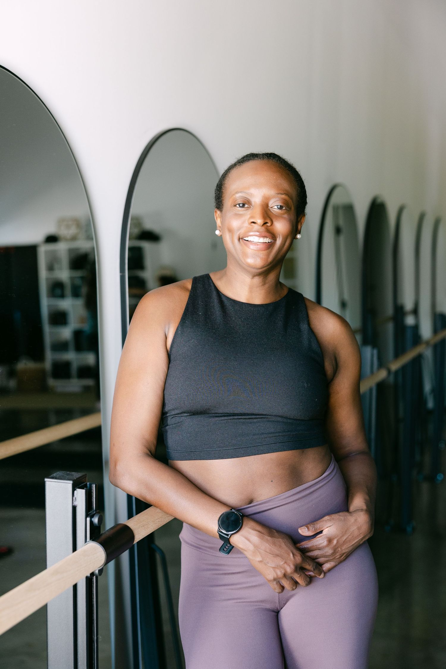 Smiling woman in black crop top and purple leggings leaning on barre in fitness studio.