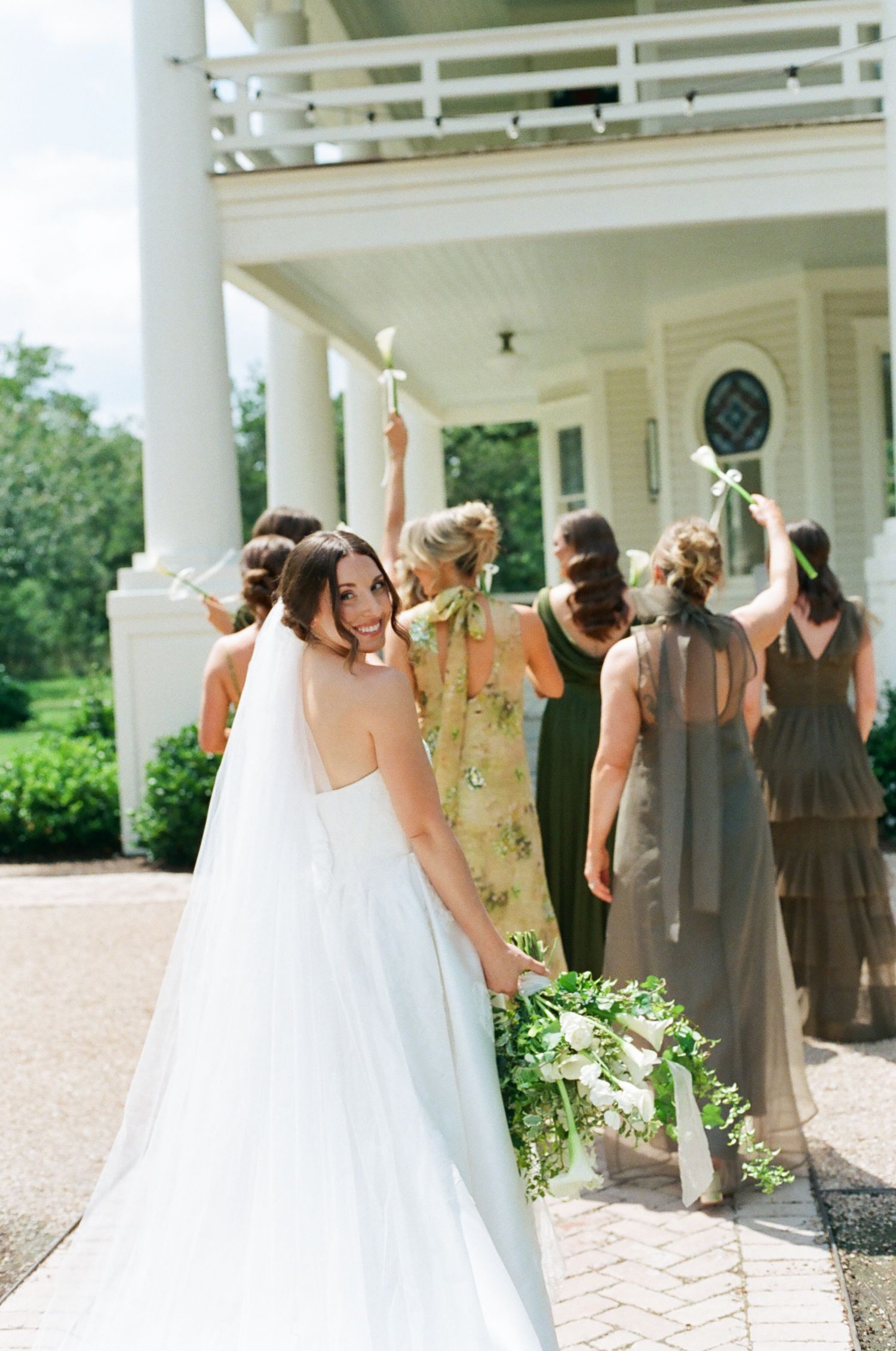 Smiling bride in white gown turns back as bridesmaids in green and taupe dresses raise flowers outside a white columned estate.