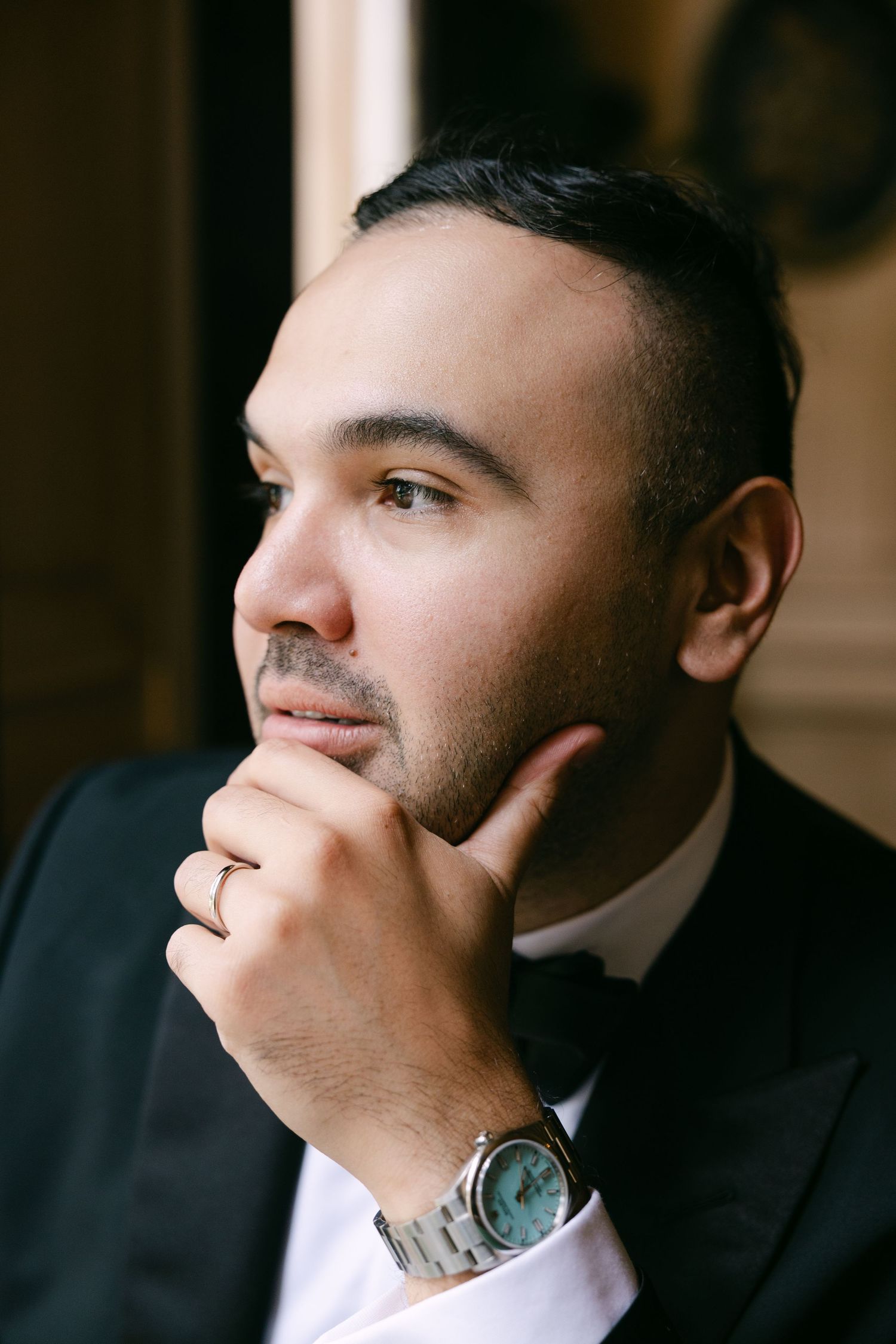 Handsome groom in a black tuxedo and bow tie poses thoughtfully with hand on chin, wearing a luxury watch.