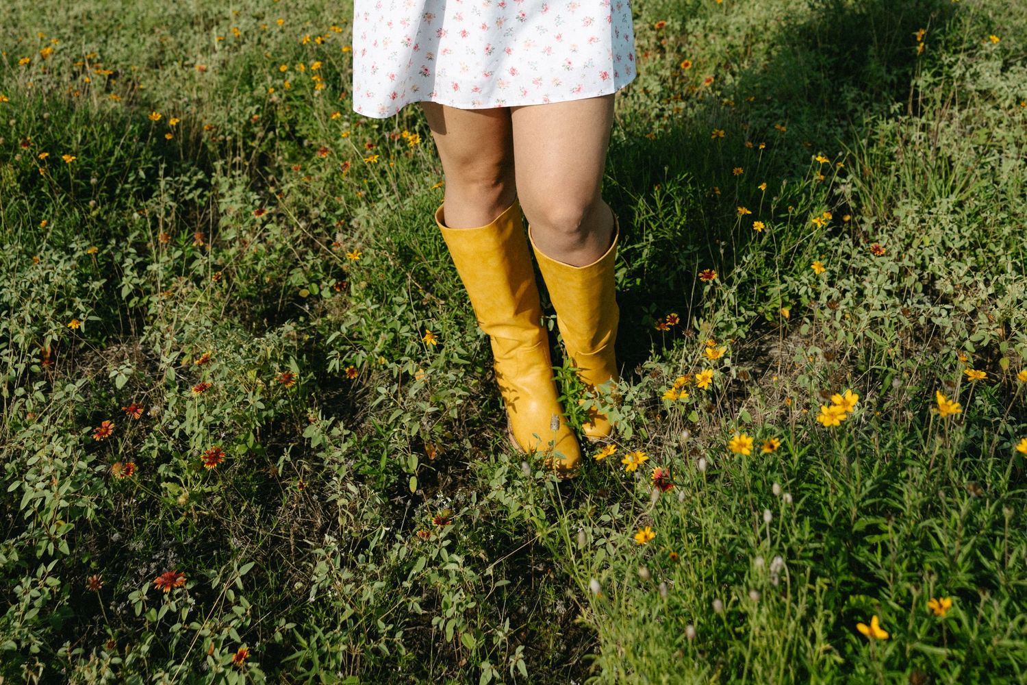 Woman in white polka dot dress wearing yellow rain boots standing in a wildflower meadow.
