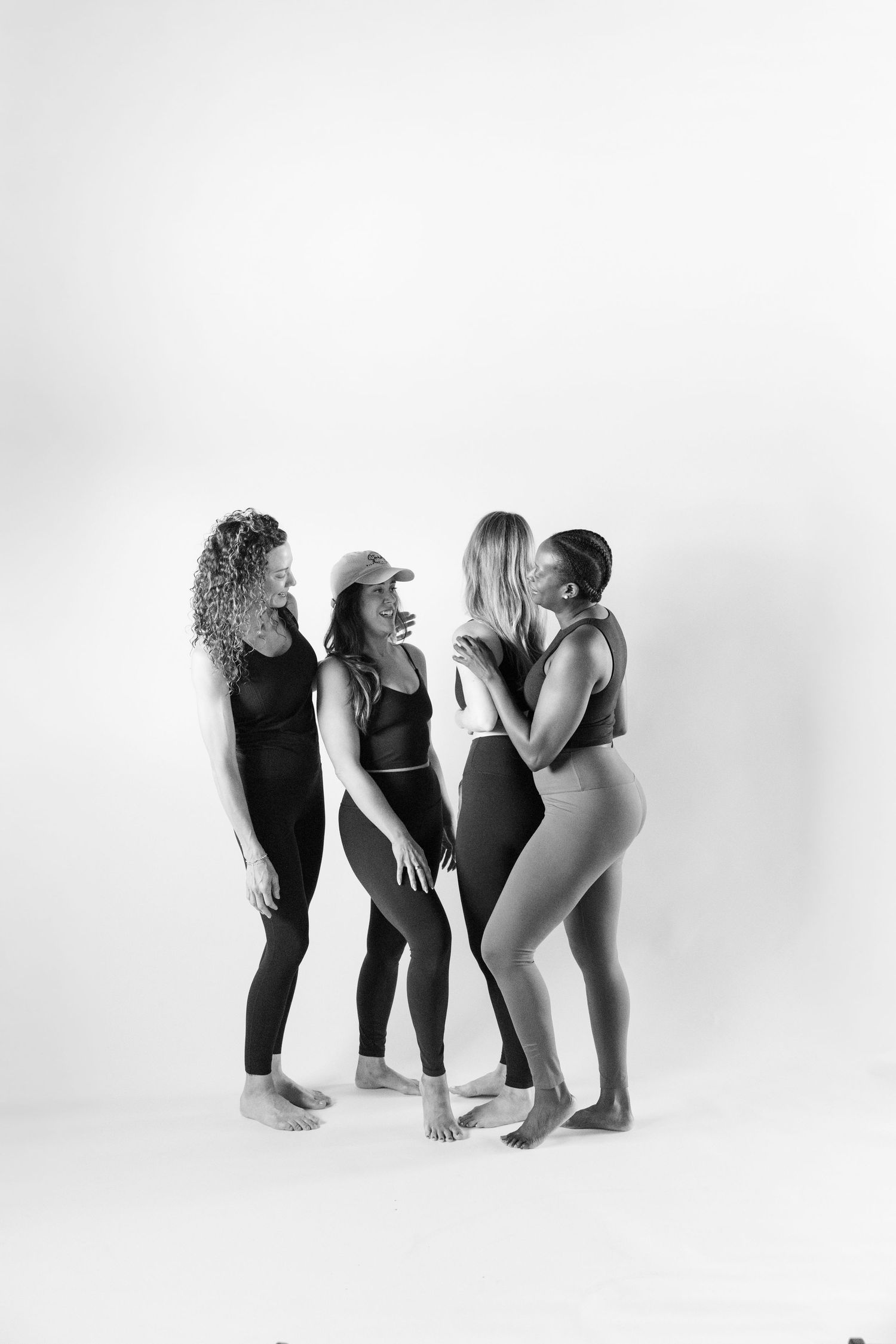 Four diverse women in black activewear laugh together in a black and white studio portrait.