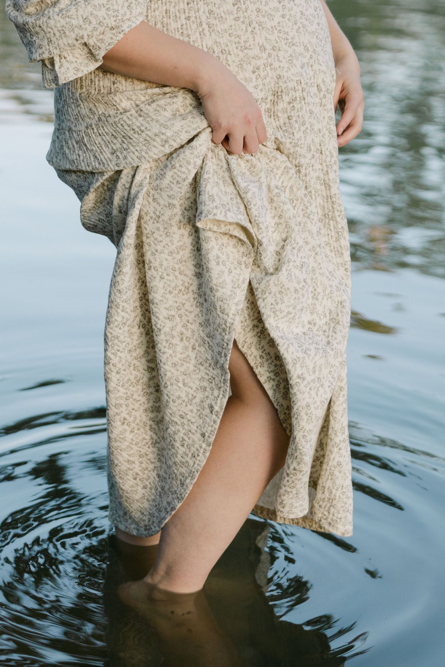 Pregnant woman in floral dress lifting skirt while standing barefoot in water.