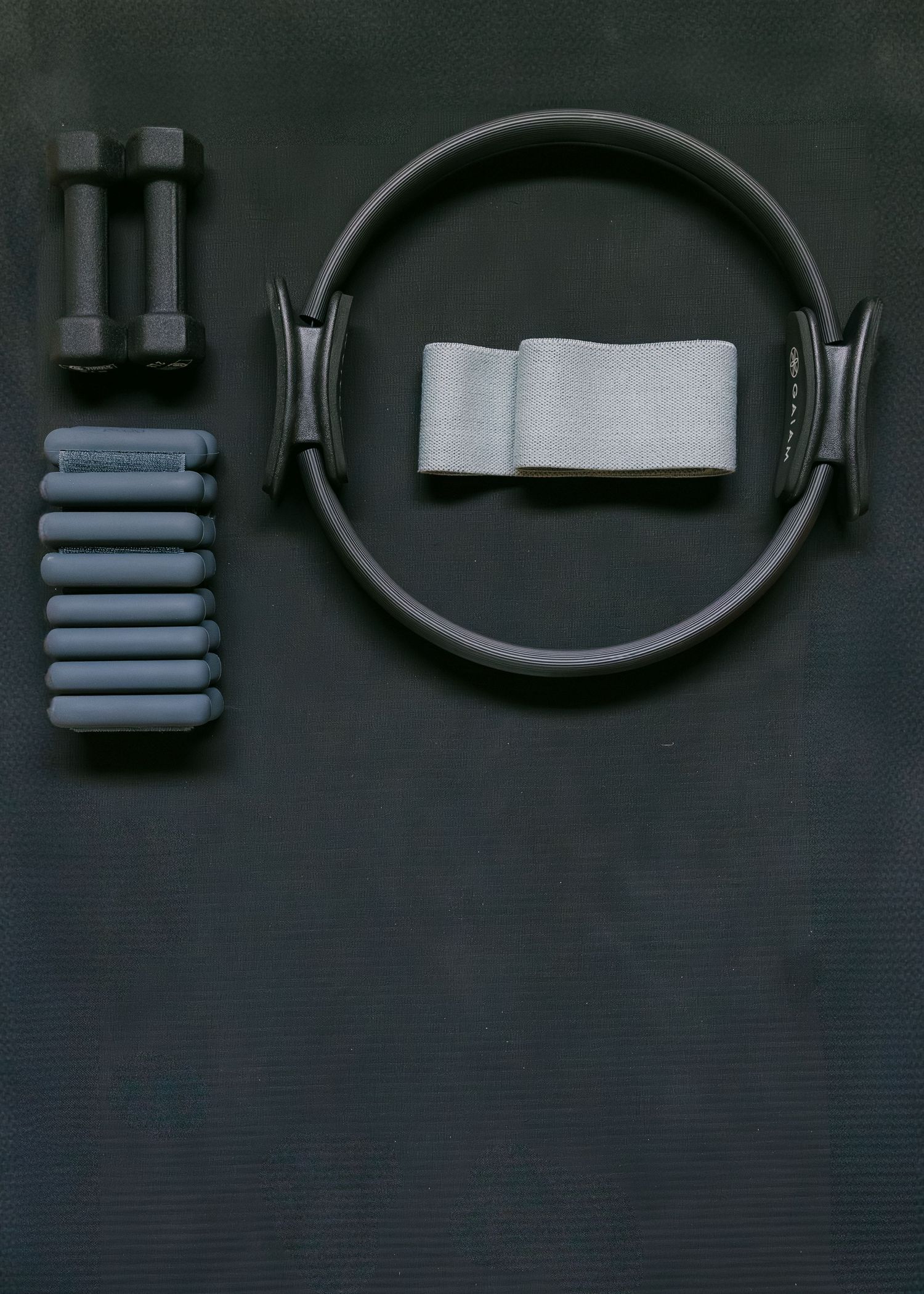 Flat lay of pilates equipment including a ring, resistance band, dumbbells, and foam roller.