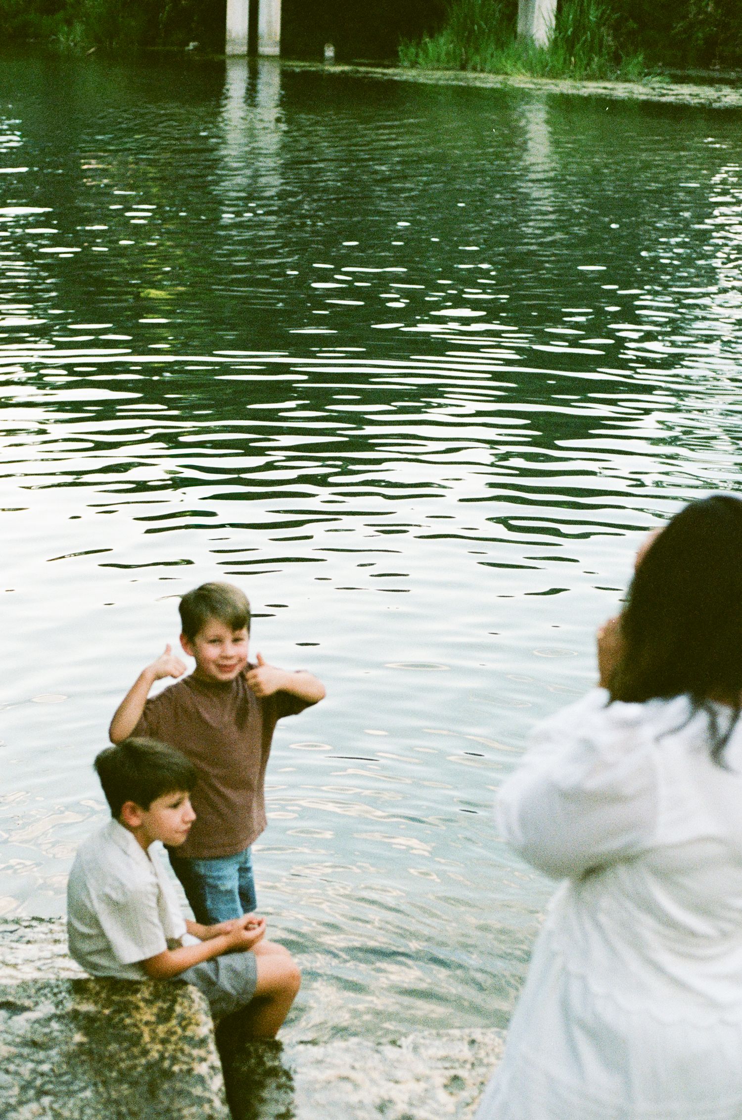 Two young boys sitting by a peaceful river's edge while a woman in white watches during a family photo session.