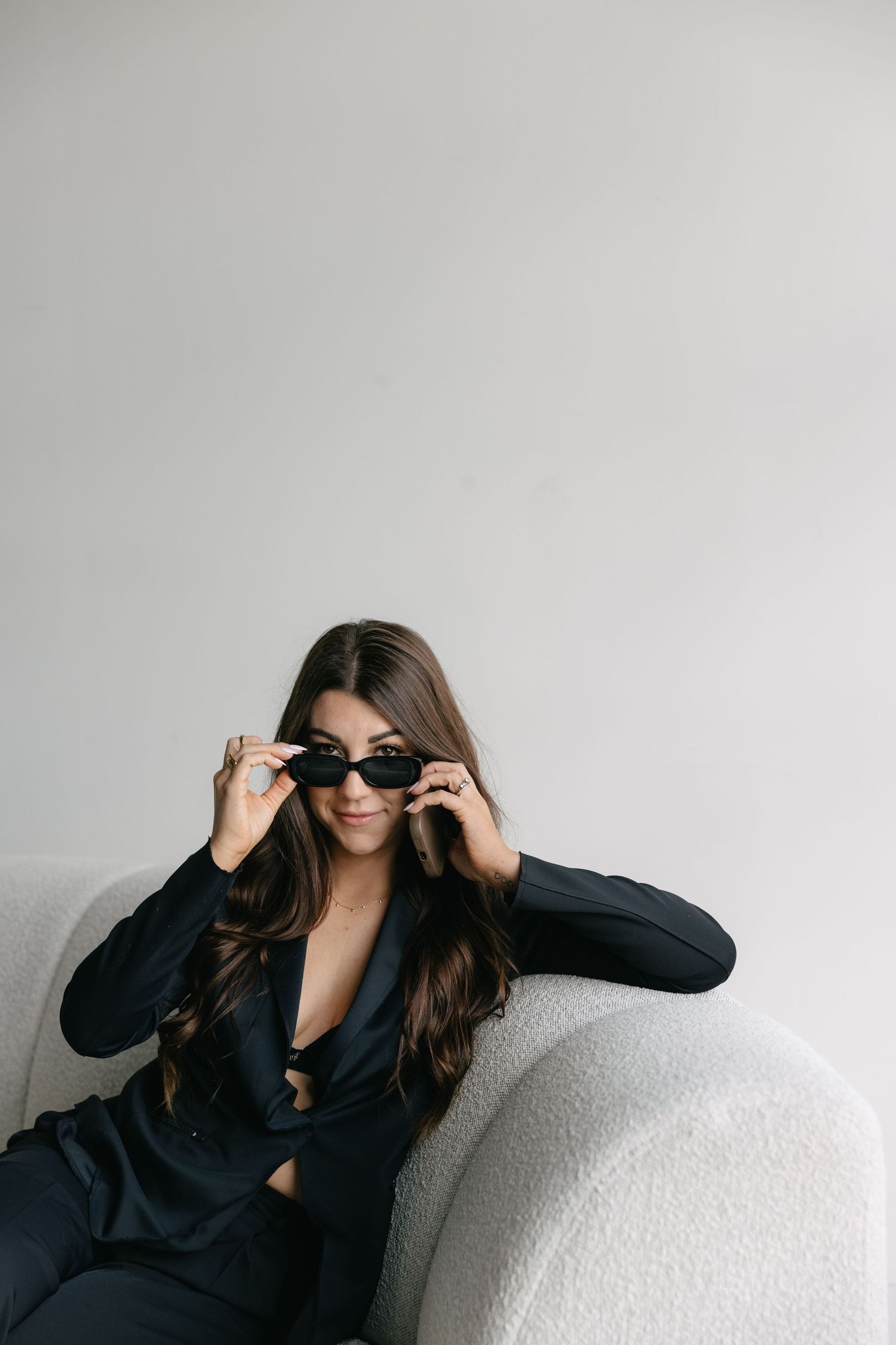 Stylish brunette woman in black suit lowering sunglasses while seated in white chair.
