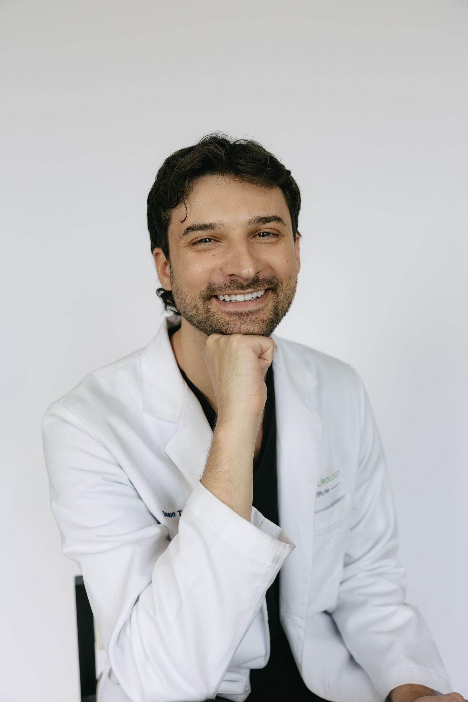 Smiling male doctor in white coat sitting relaxed against white background.