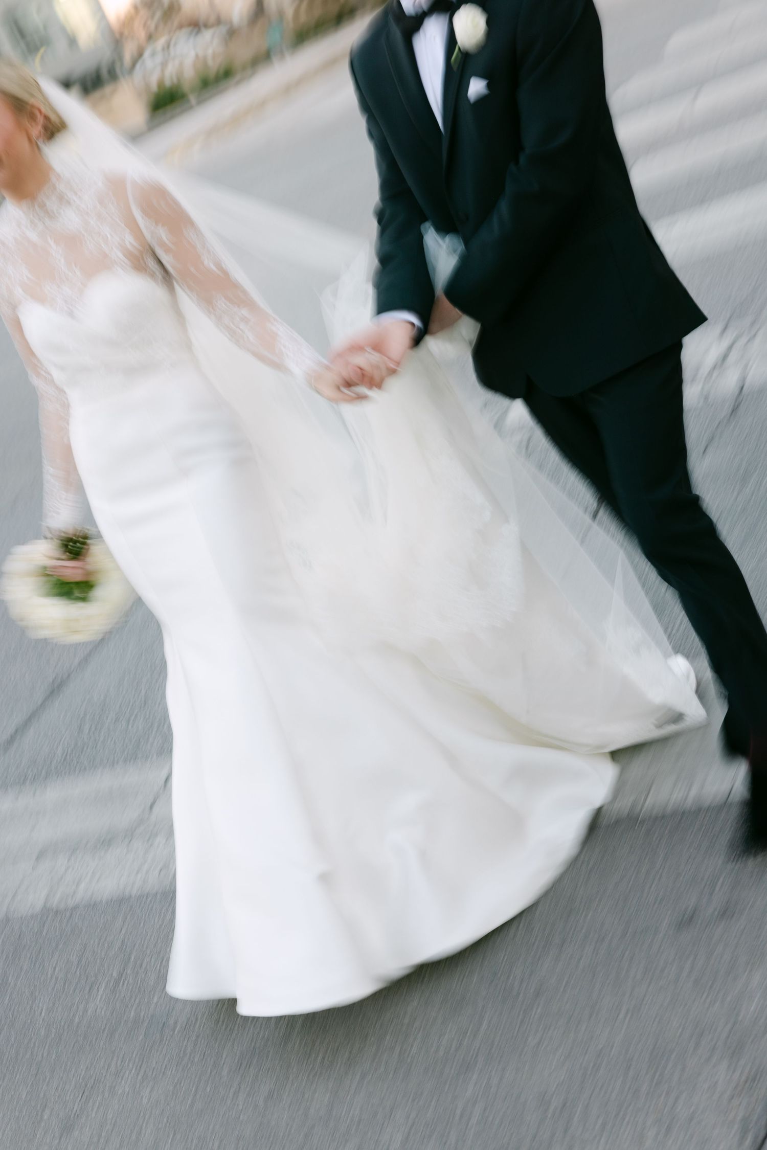 Bride in white mermaid gown and groom in black tuxedo walk hand in hand, dress flowing in motion blur.