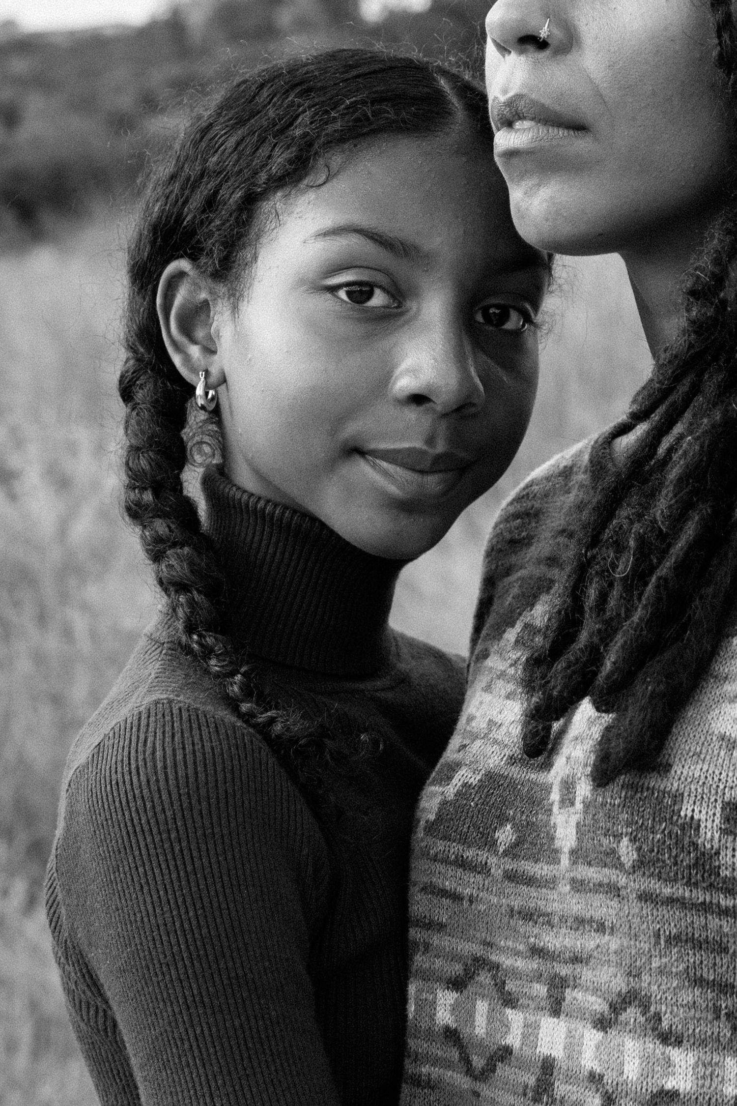 Black and white portrait of a young girl with braids leaning against a woman outdoors.
