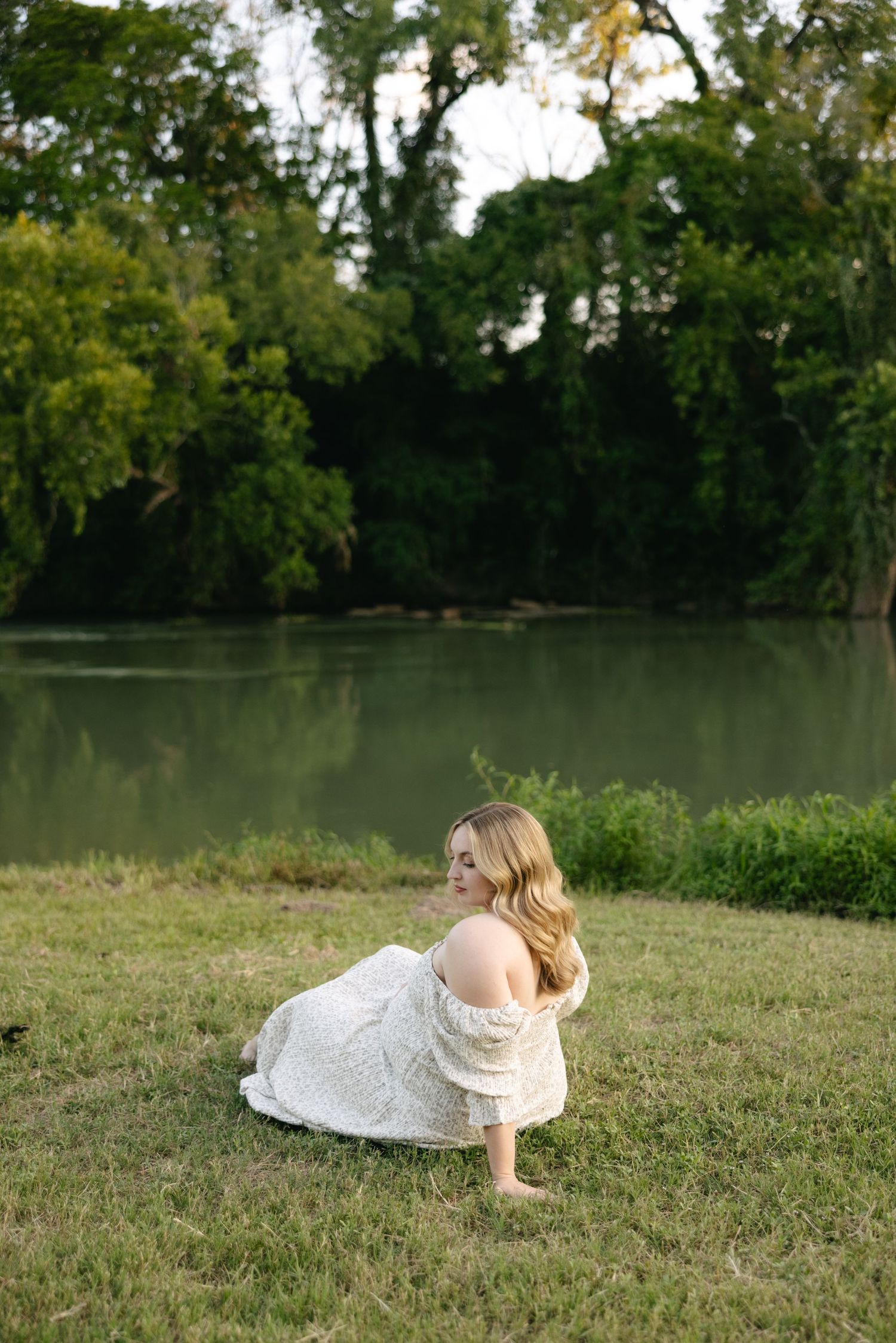 Pregnant woman in white lace dress sitting barefoot on grass by serene river.