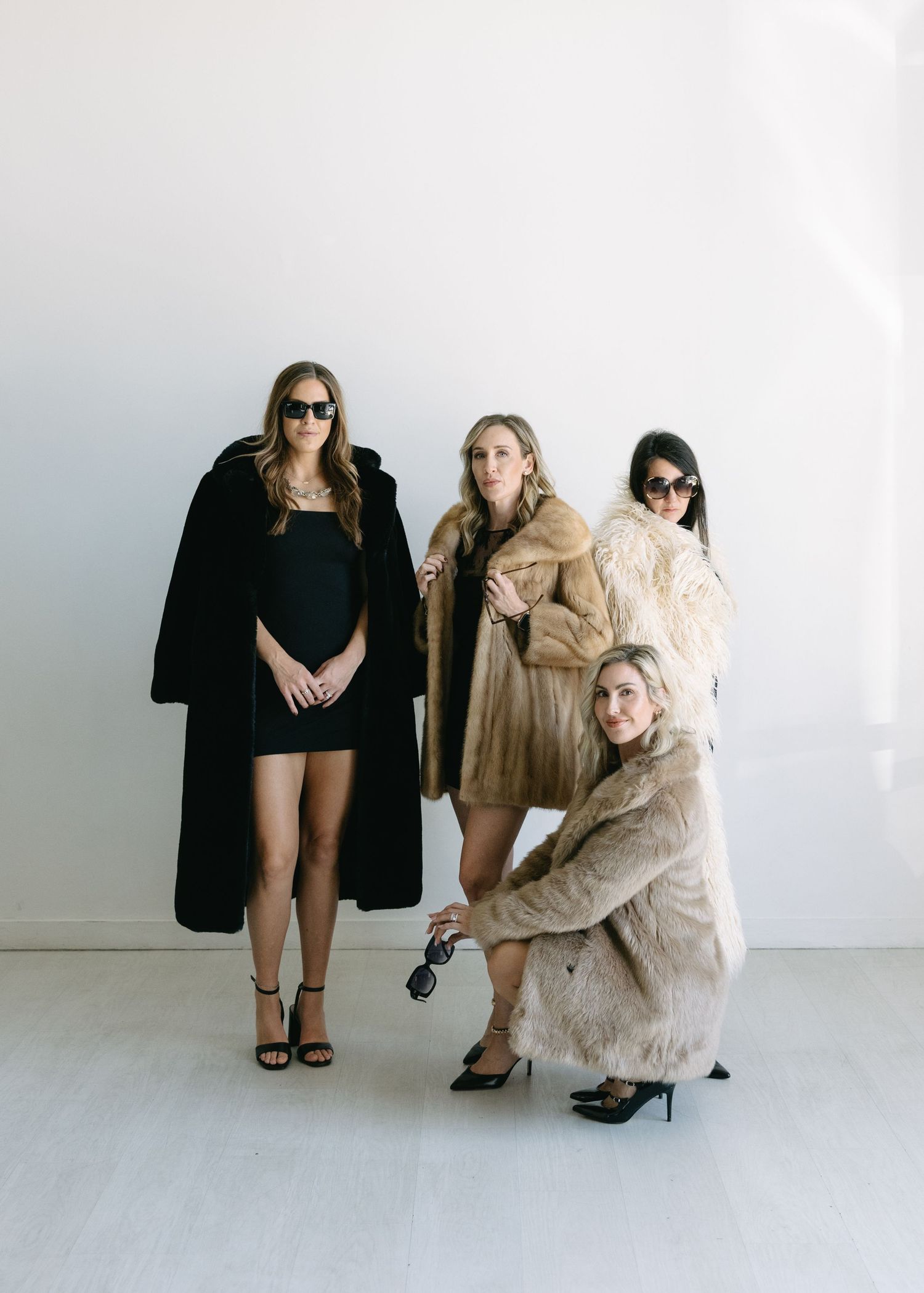 Four stylish women posing in luxury fur coats against white wall in fashion editorial shoot.