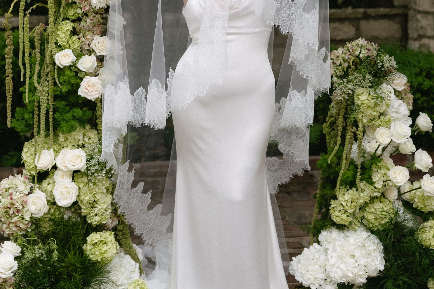 Back view of a bride in a sleek white gown with lace veil standing before a floral arch.