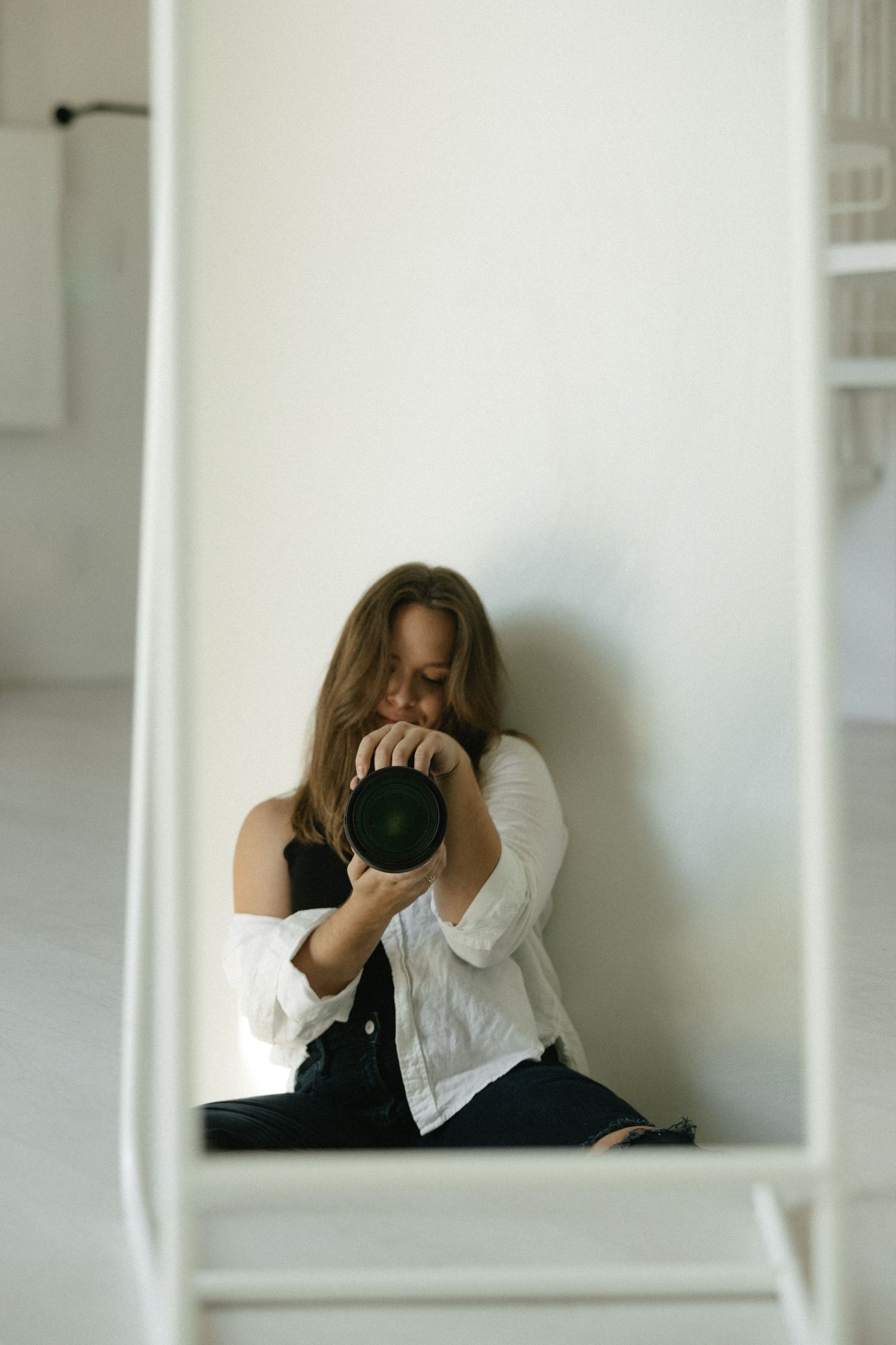 Woman photographer taking a self-portrait with DSLR camera while sitting against white wall.