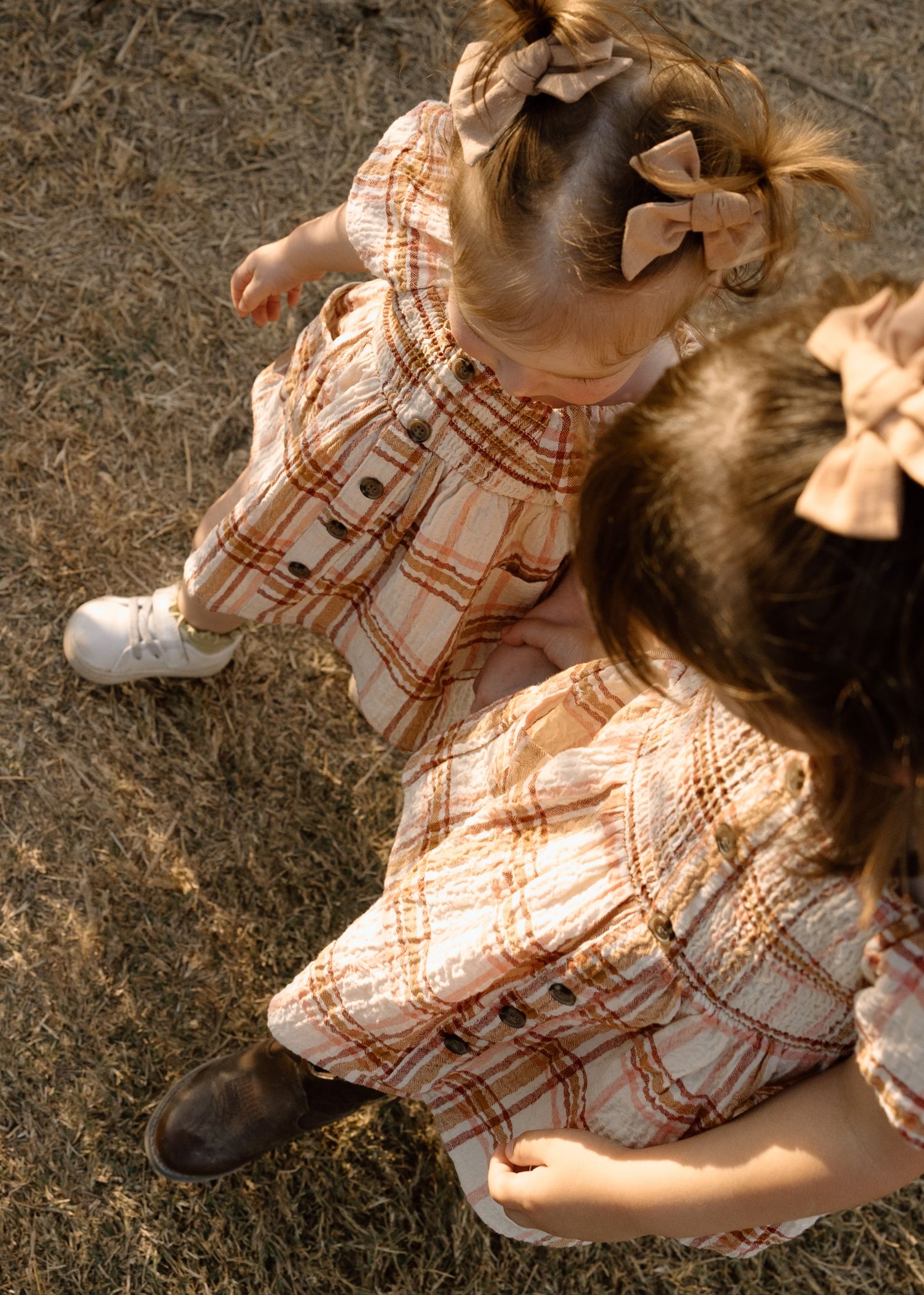 Two young girls in matching plaid dresses with bow hair clips sitting together on dry grass.