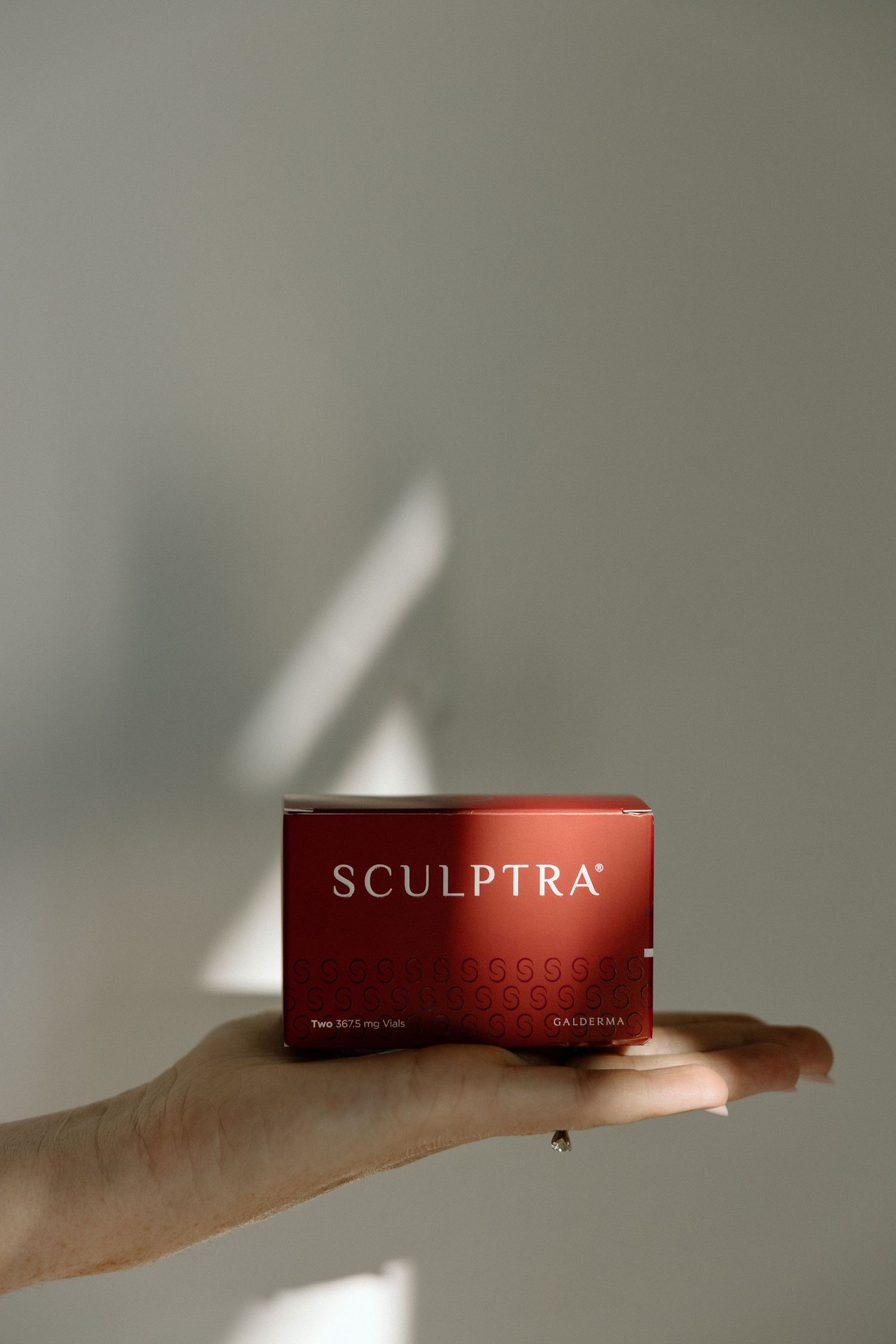 Hand holding red Sculptra collagen stimulator box against neutral gray background.