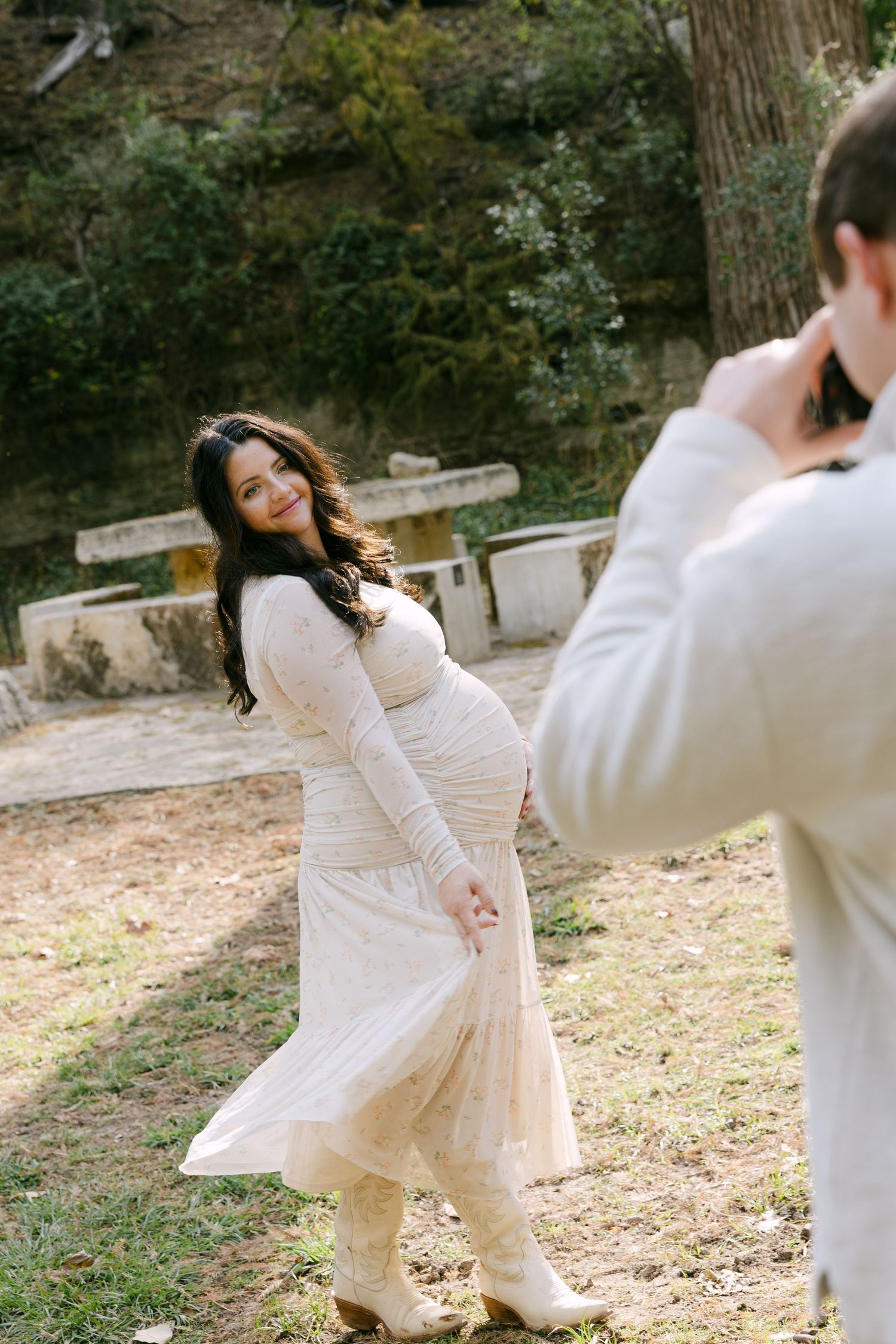 Smiling pregnant woman in white lace dress posing while partner photographs her outdoors.