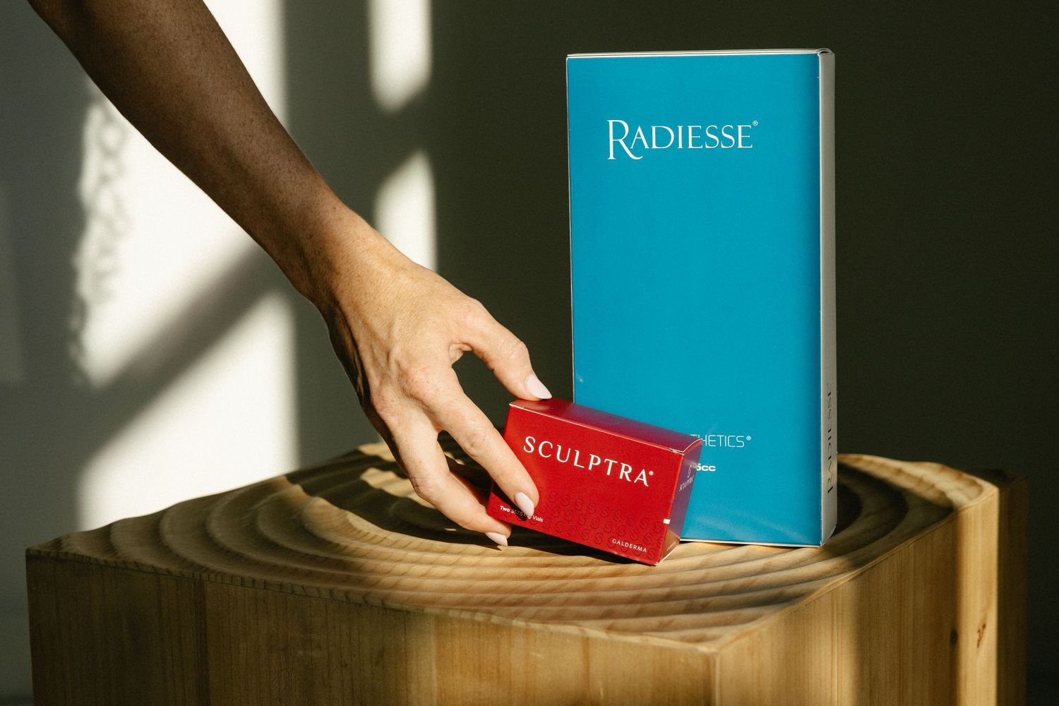 Hand reaching for Sculptra and Radiesse dermal filler boxes on a wooden pedestal table.