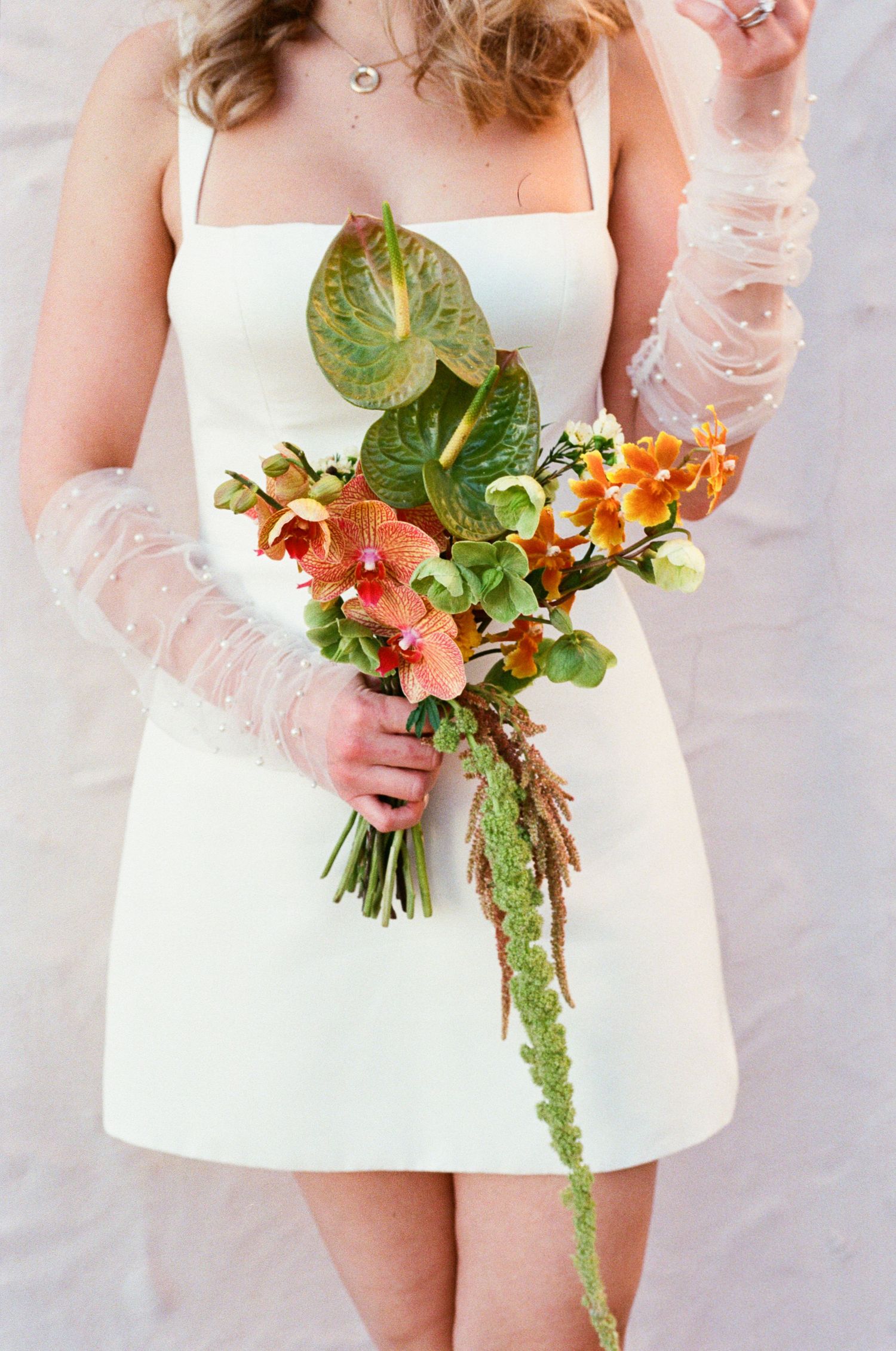 Bride in white mini dress holding a tropical orange and green bridal bouquet with sheer embellished gloves.