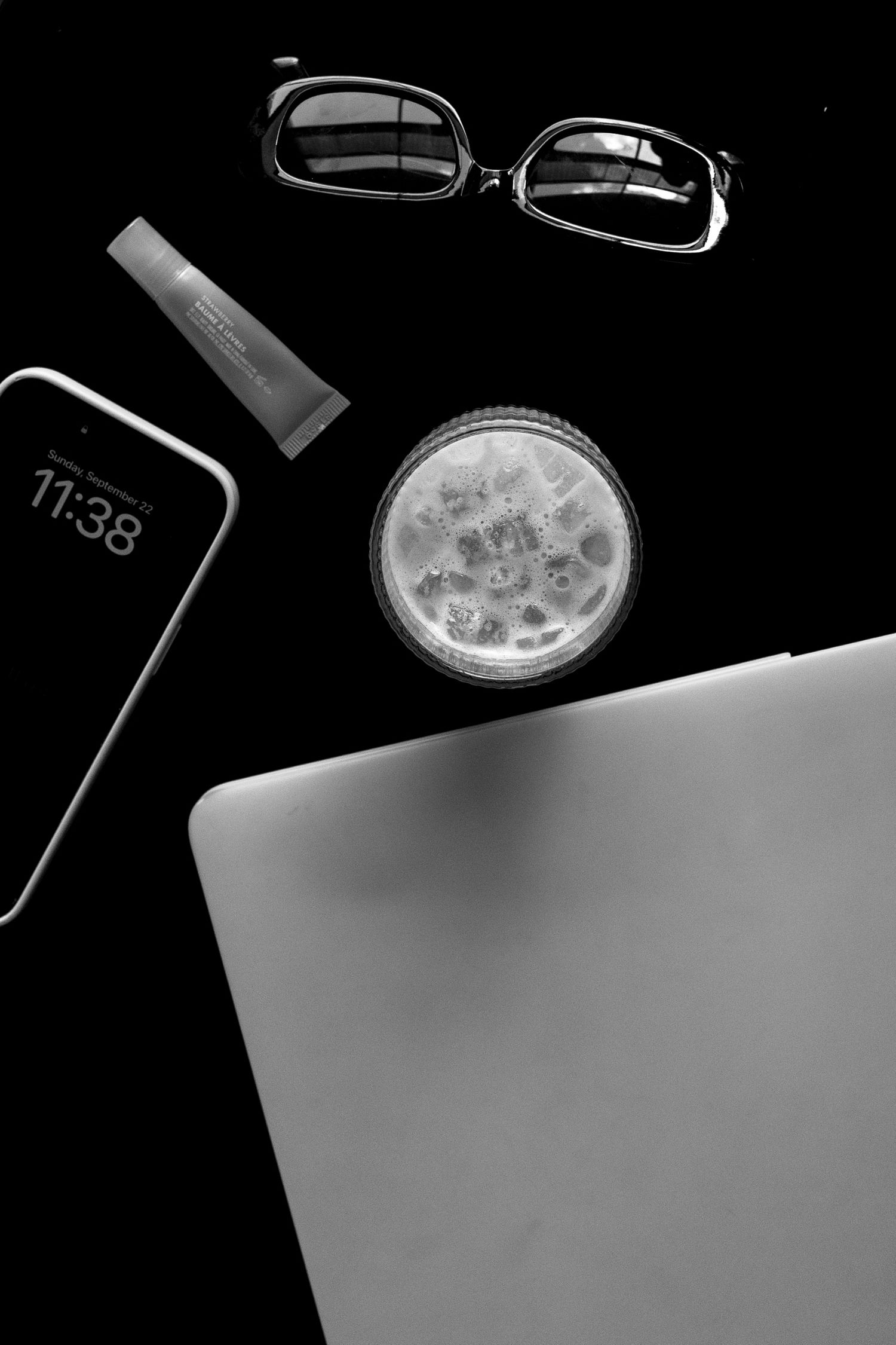 Black and white flat lay of laptop, smartphone showing 11:38, glasses, and drink.