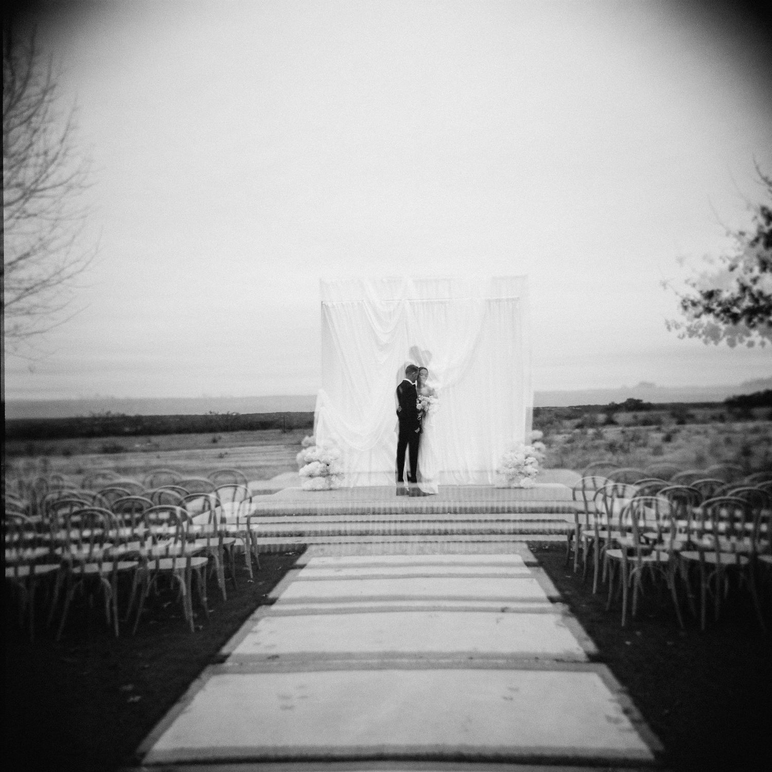Black and white photo of couple kissing at outdoor ceremony altar with empty chairs.