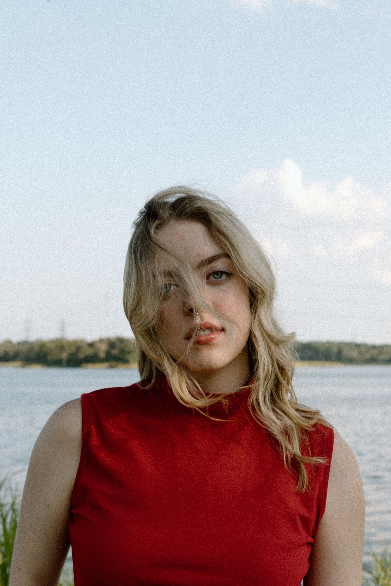 Young blonde woman in red sleeveless top with hair blowing across face, standing near a lake under blue sky.
