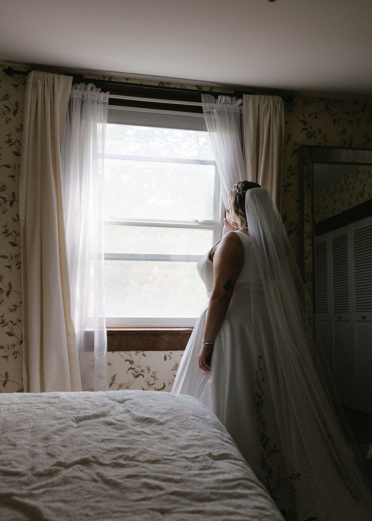 Bride in a white gown and veil gazes out a bedroom window on her wedding day.