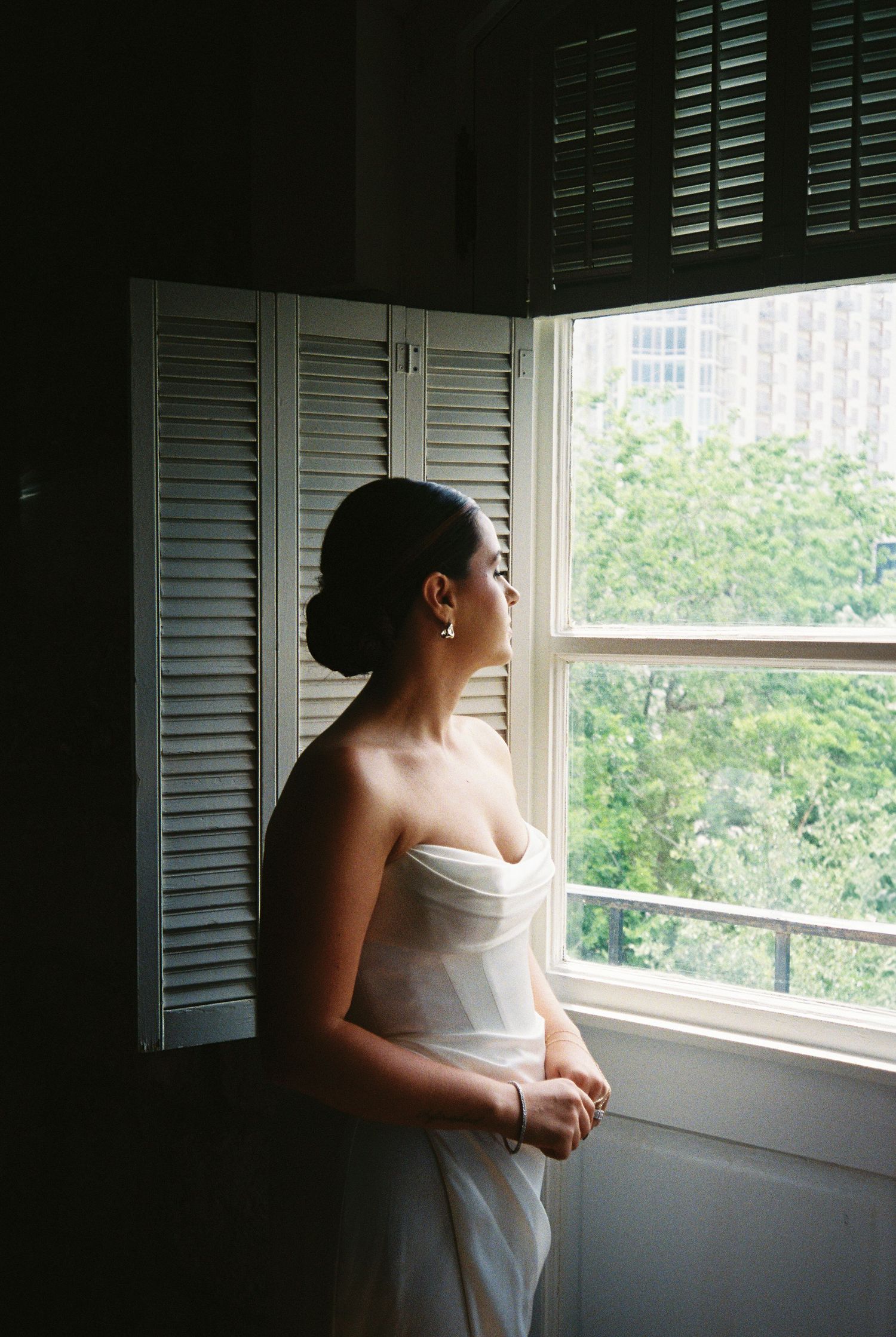 Bride in strapless white gown gazes out a window with wooden shutters in soft light.