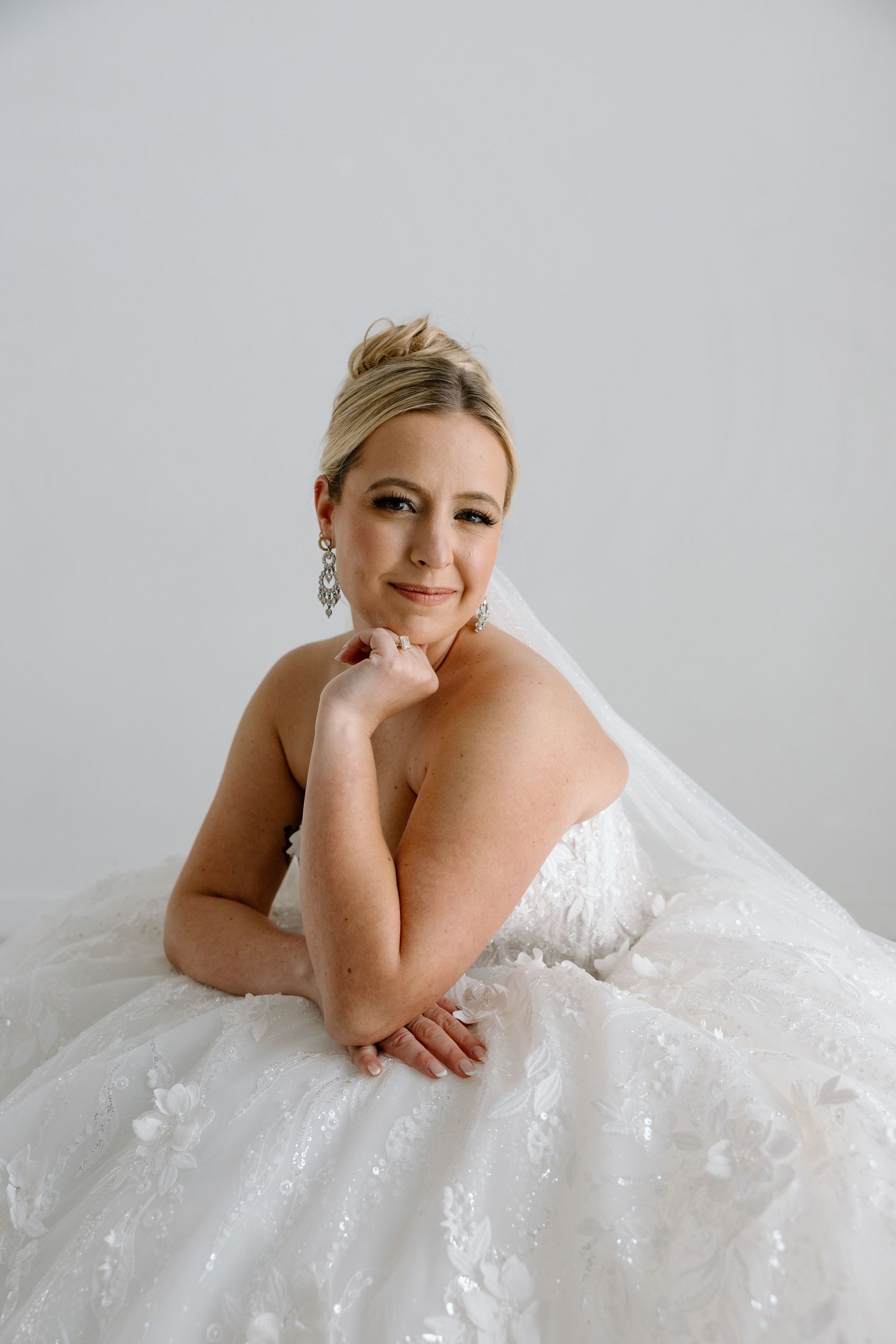 Elegant blonde bride in a ballgown with floral lace detail posing on a white studio floor.