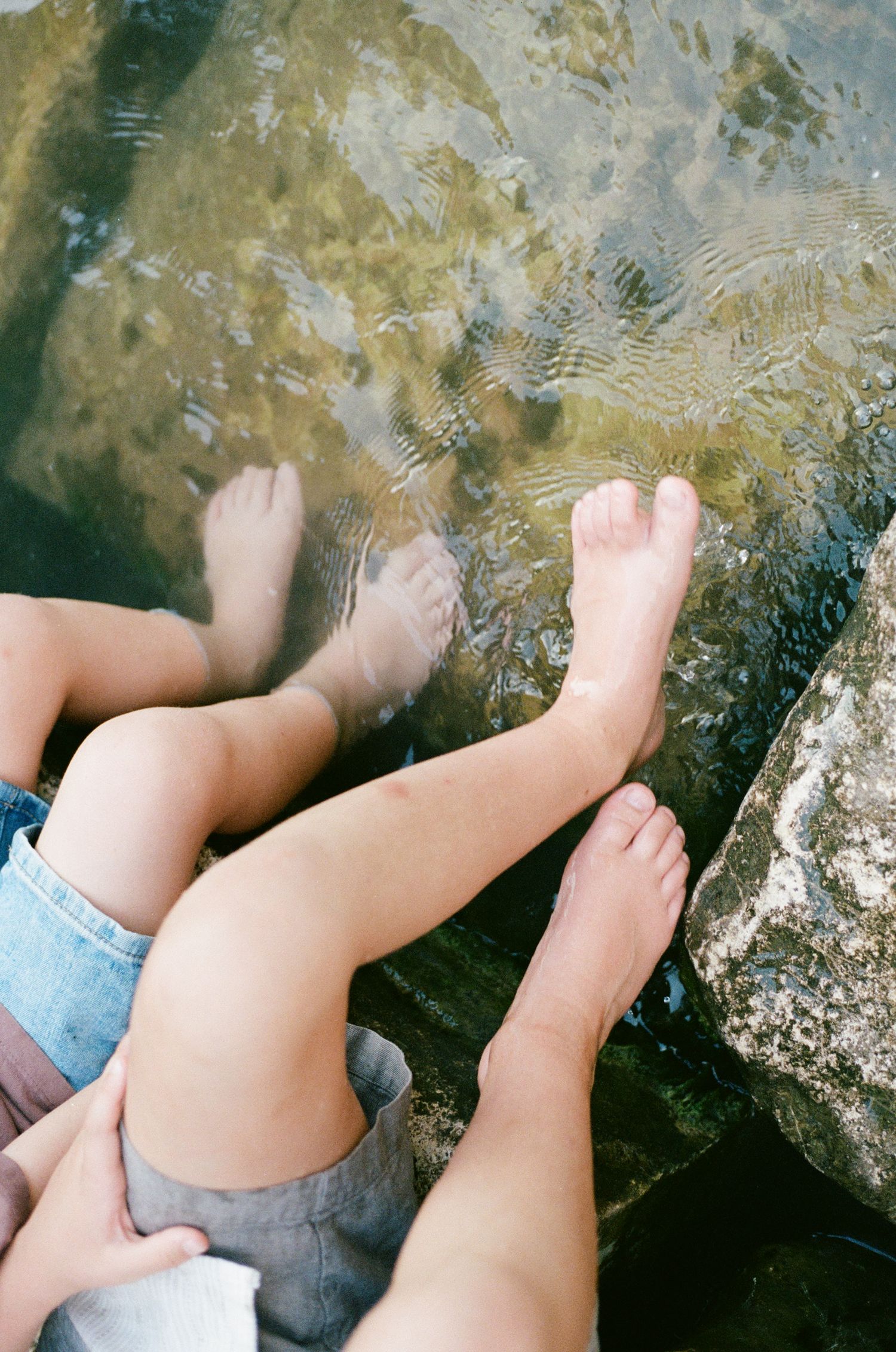 Close-up of children's bare feet dangling in clear shallow water over rocky riverbed during family photos.
