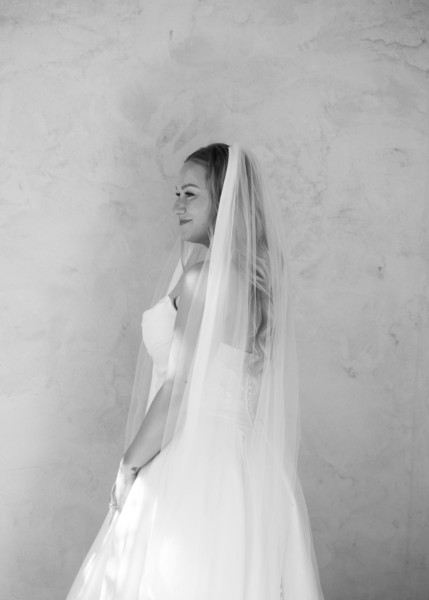 Elegant bride in a white gown and long veil poses gracefully in a black and white bridal portrait.
