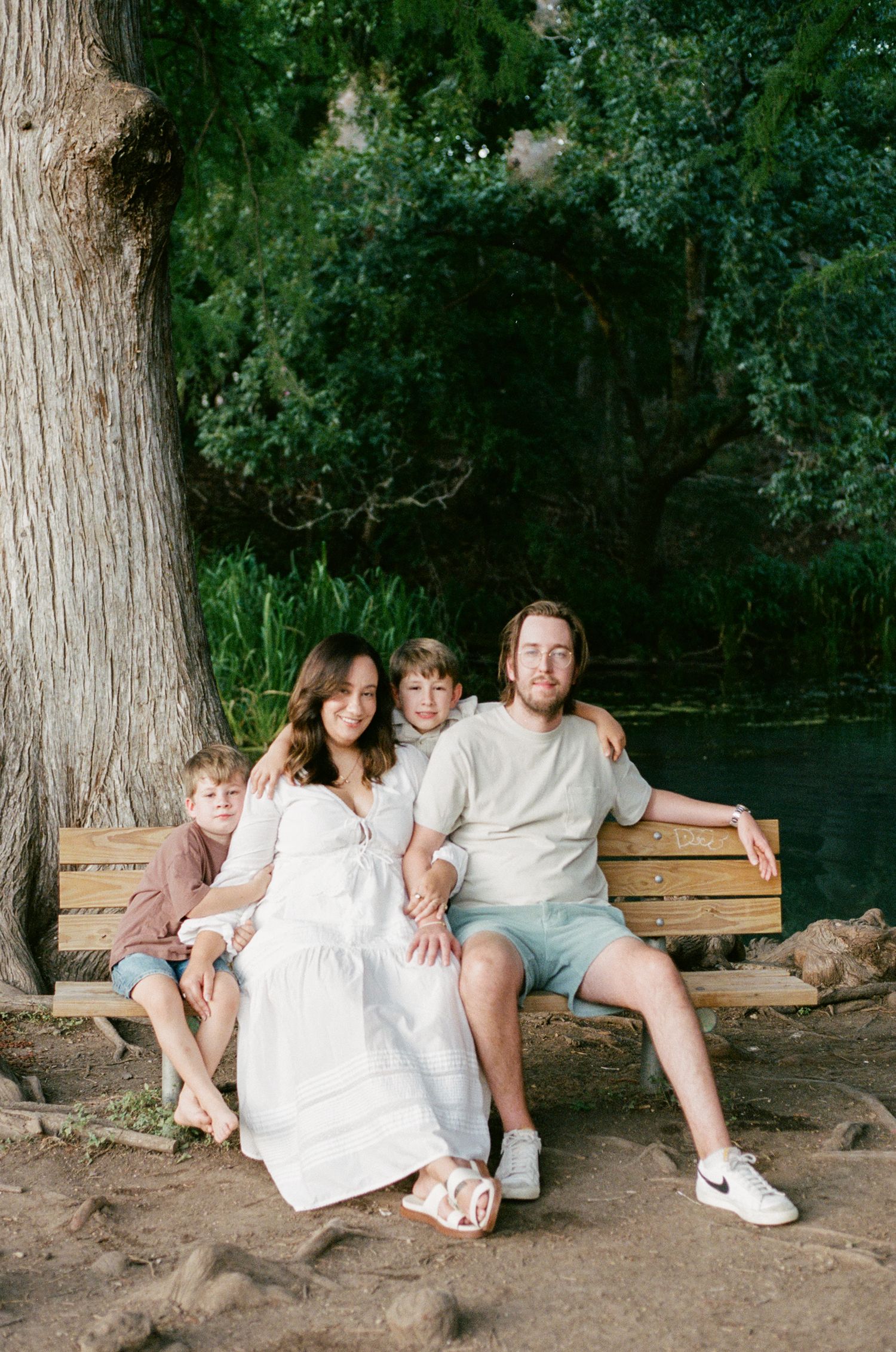 Happy family of four sits together on a wooden bench beside a tranquil green river surrounded by lush trees.