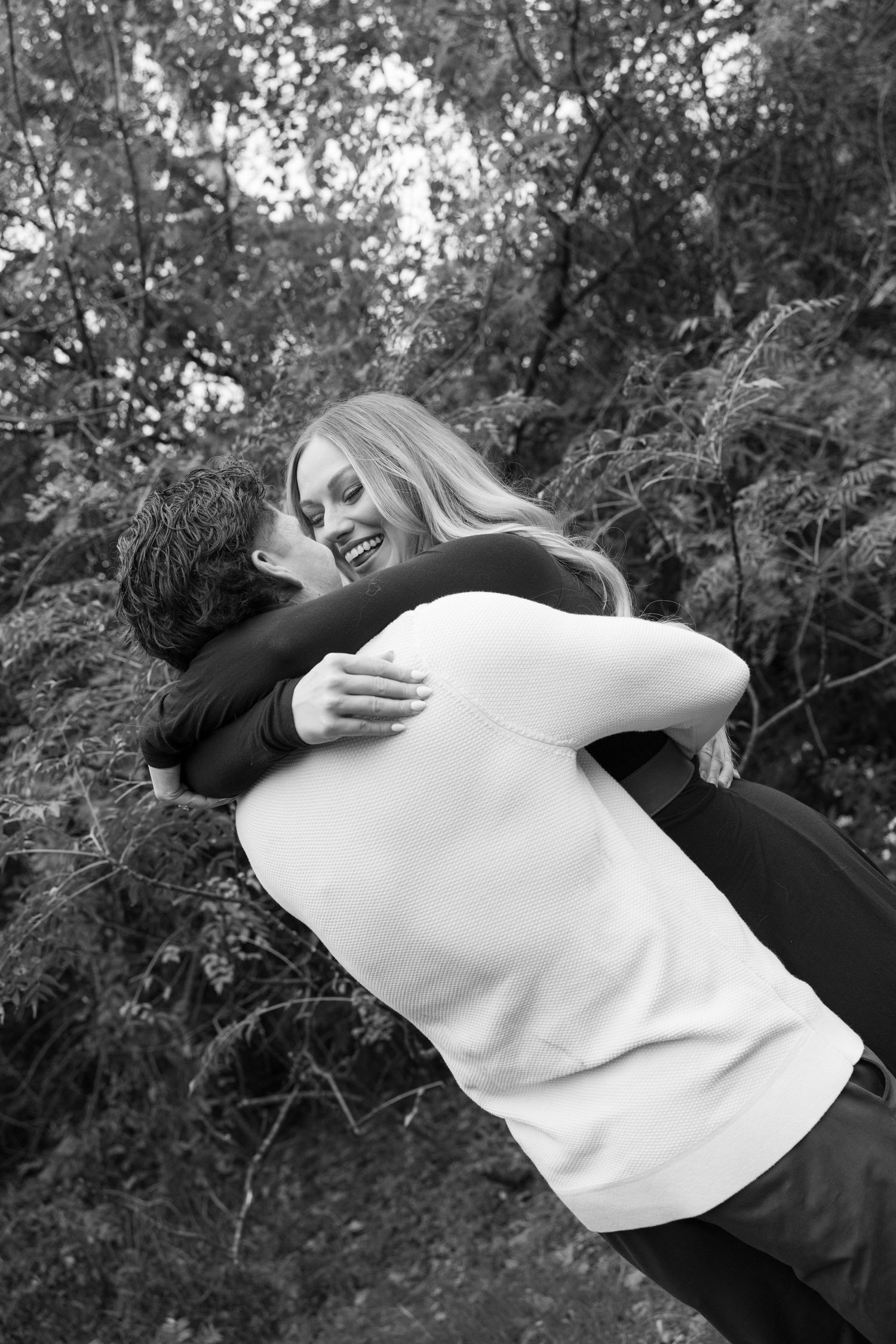 Black and white photo of a couple embracing and laughing outdoors among lush green trees.