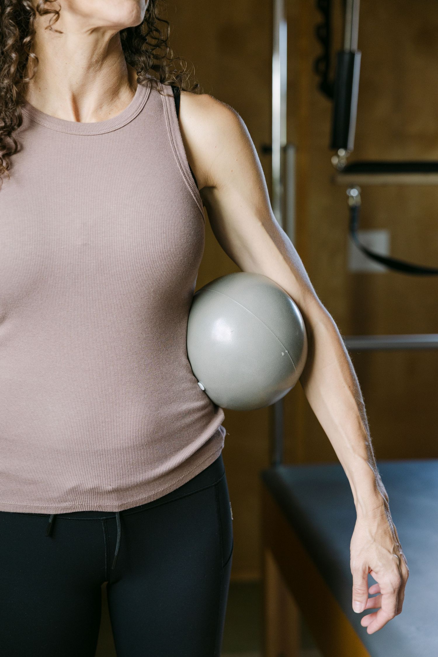 Woman in a tank top holding a small pilates ball against her side in a gym.