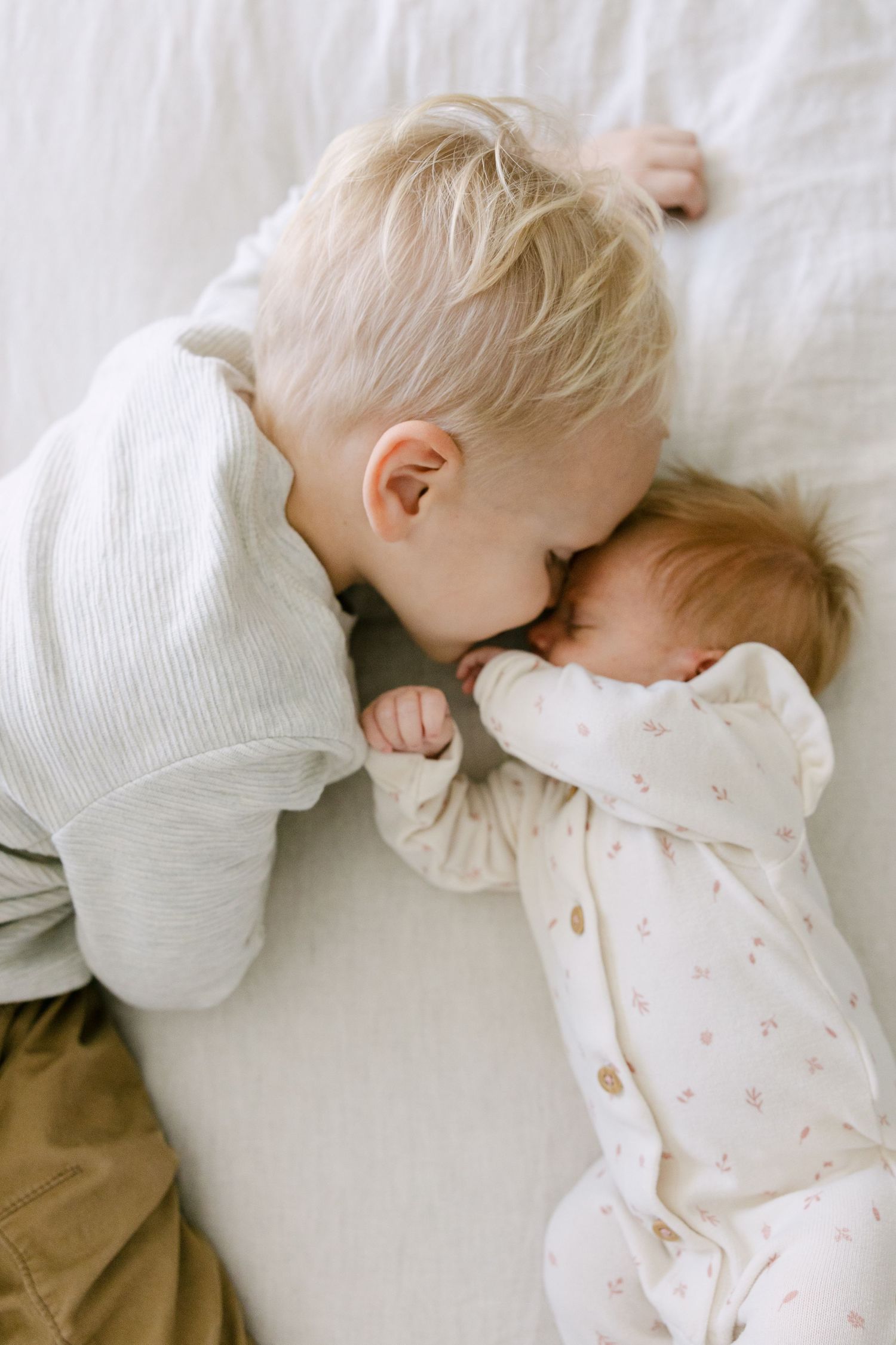 Toddler boy gently kissing sleeping newborn baby sibling on white bedding.