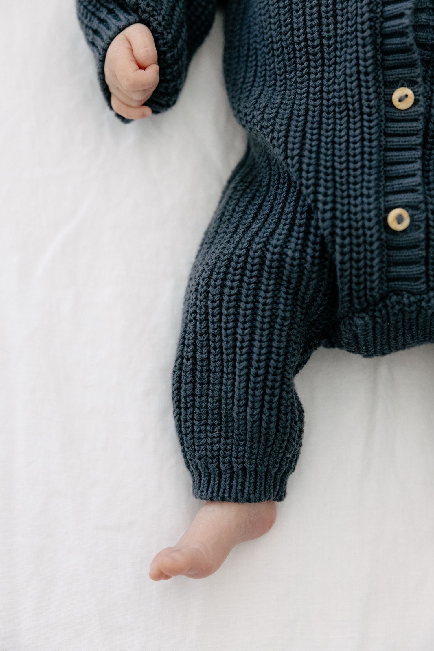 Close-up of a newborn baby wearing a chunky dark teal knit romper lying on white fabric.