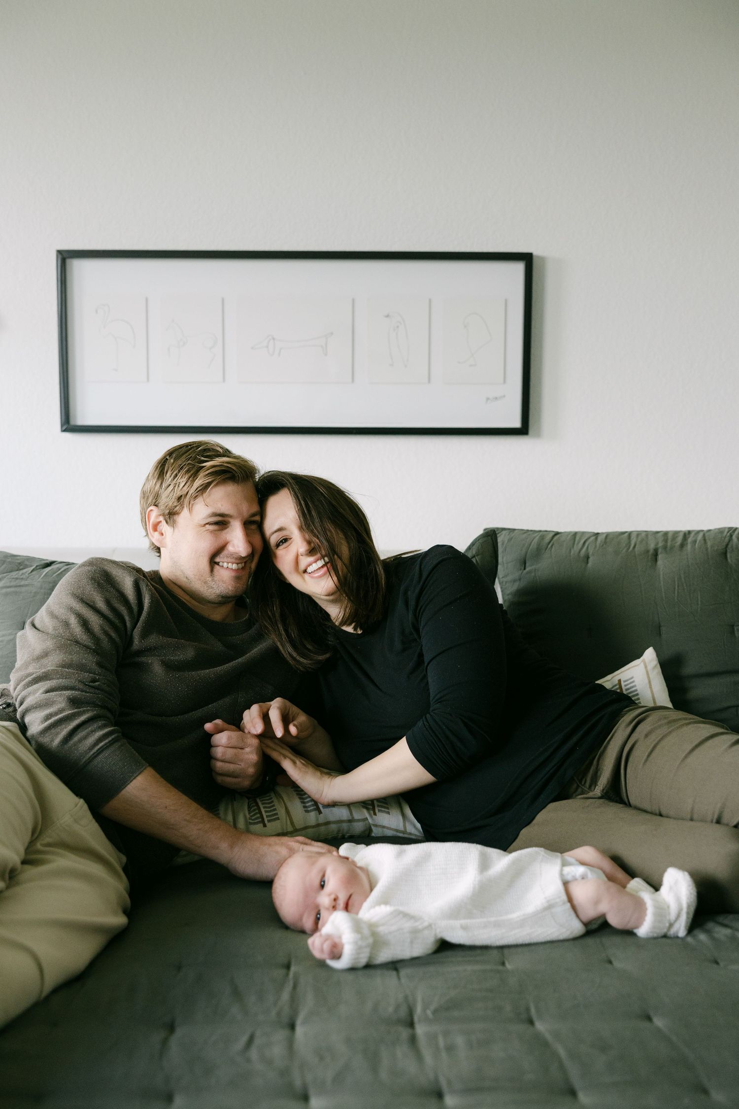 Happy couple laughing together on a green sofa with their sleeping newborn baby in white.
