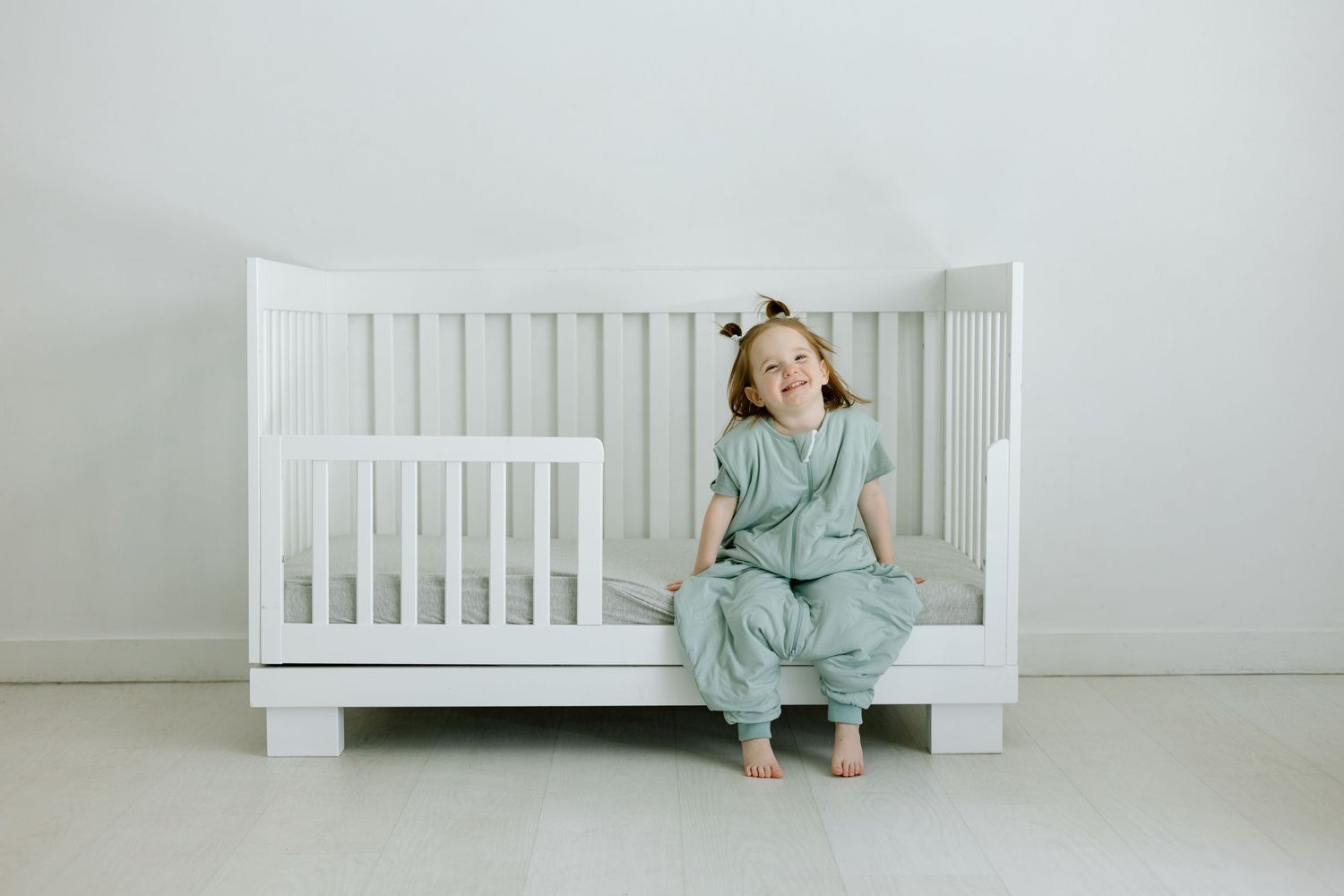 Young girl in sage green romper sitting on white convertible crib in bright nursery room.
