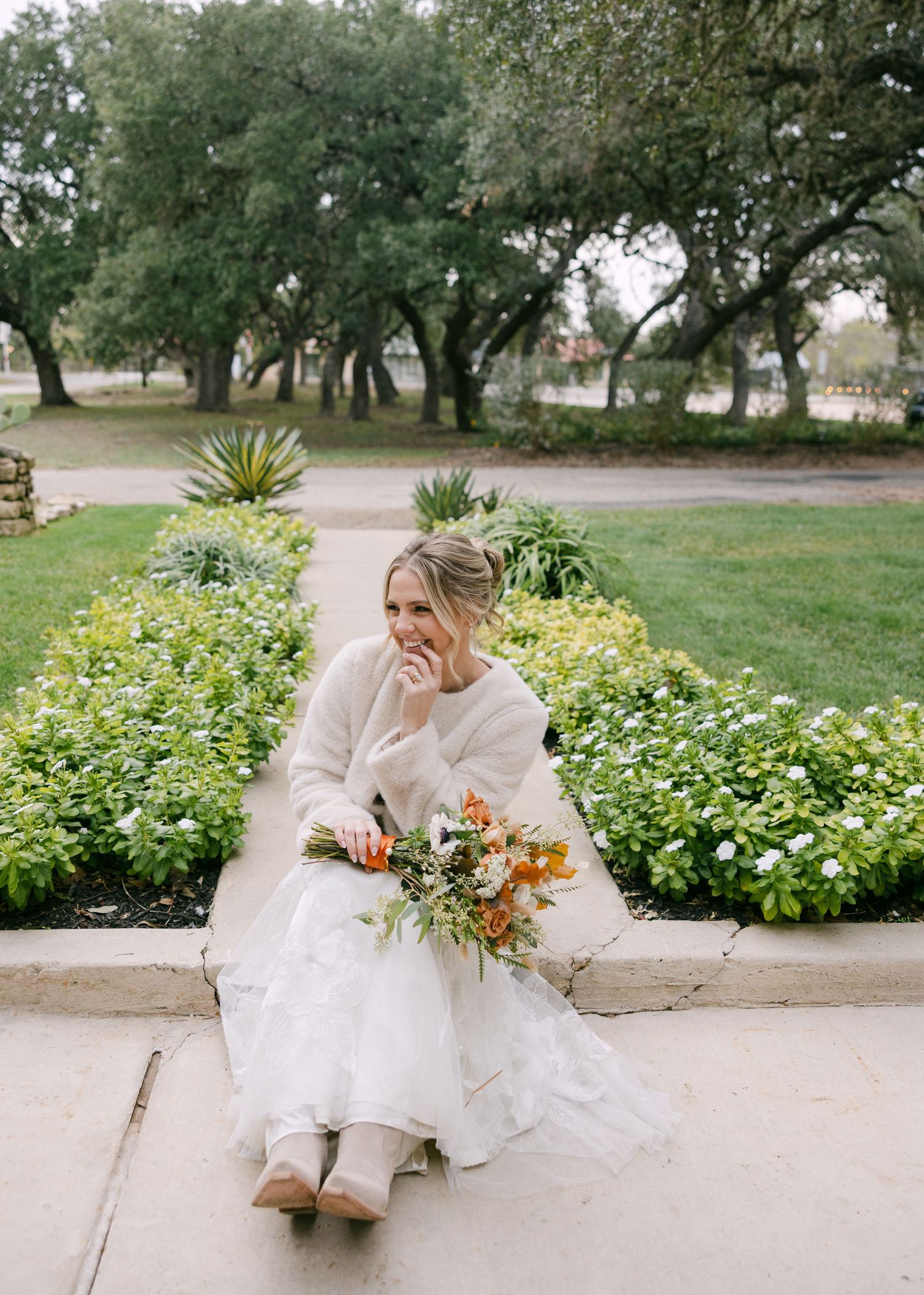Joyful bride in a white gown and cozy sweater sitting on a garden path holding a bouquet.