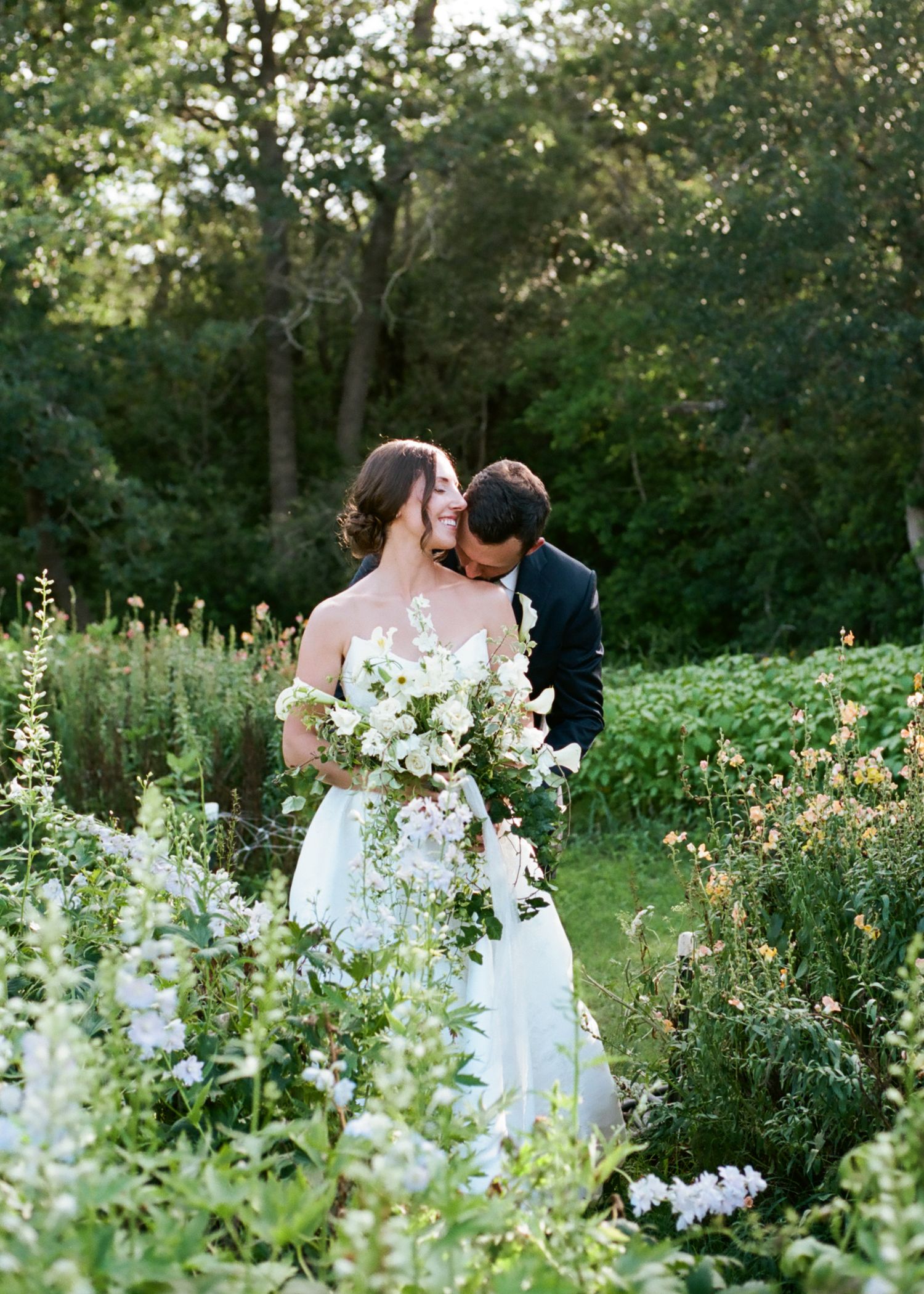 Bride and groom share a romantic moment surrounded by lush wildflowers and greenery in a garden at golden hour.