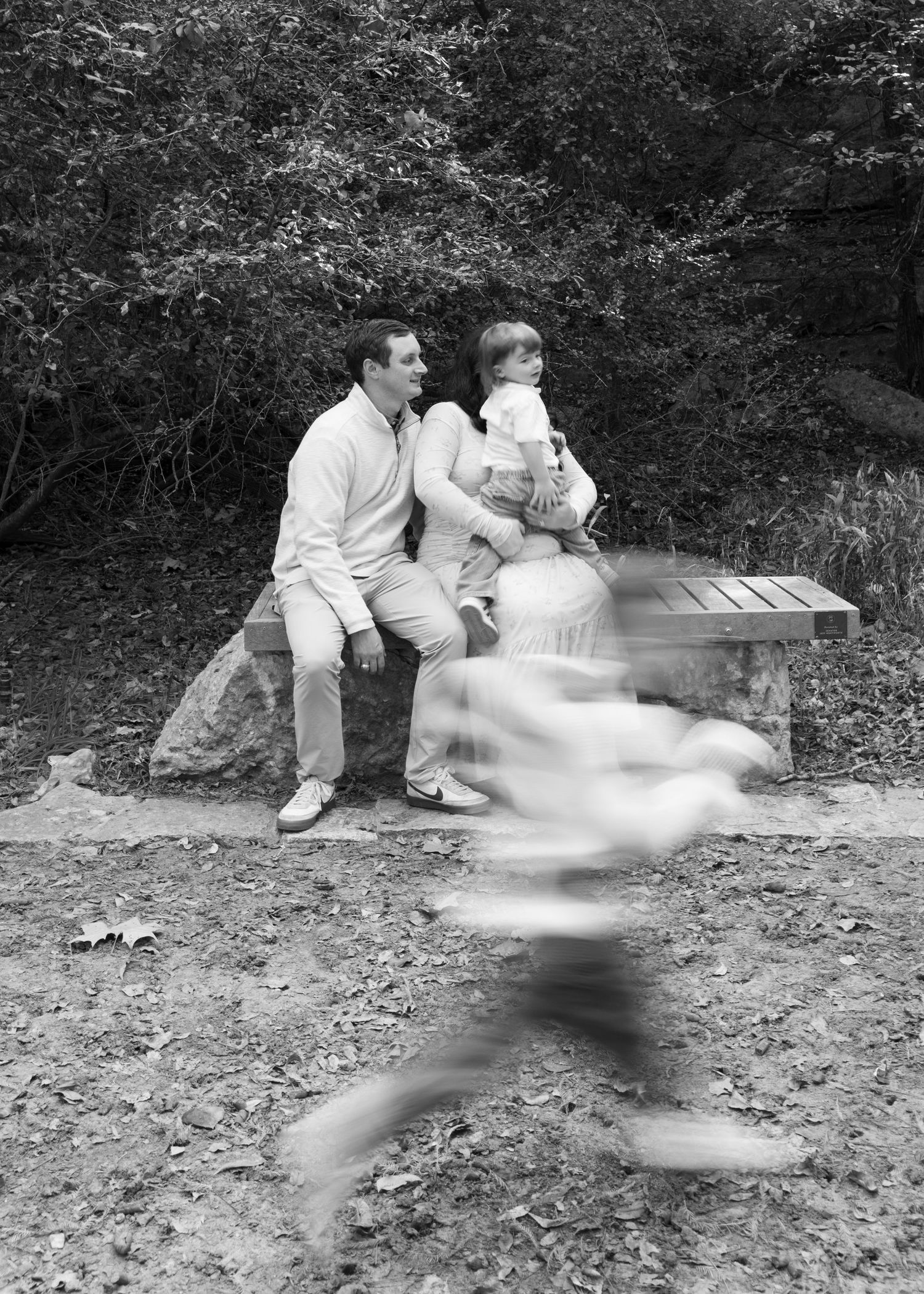 Black and white photo of a man and baby on a bench while a child runs by in a blur.