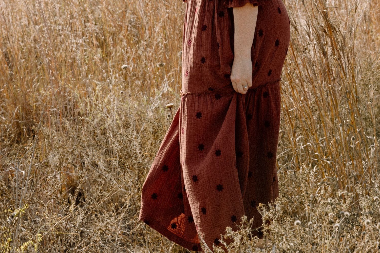 Woman in rust-colored polka dot maxi dress stands gracefully in a golden dry grass field.