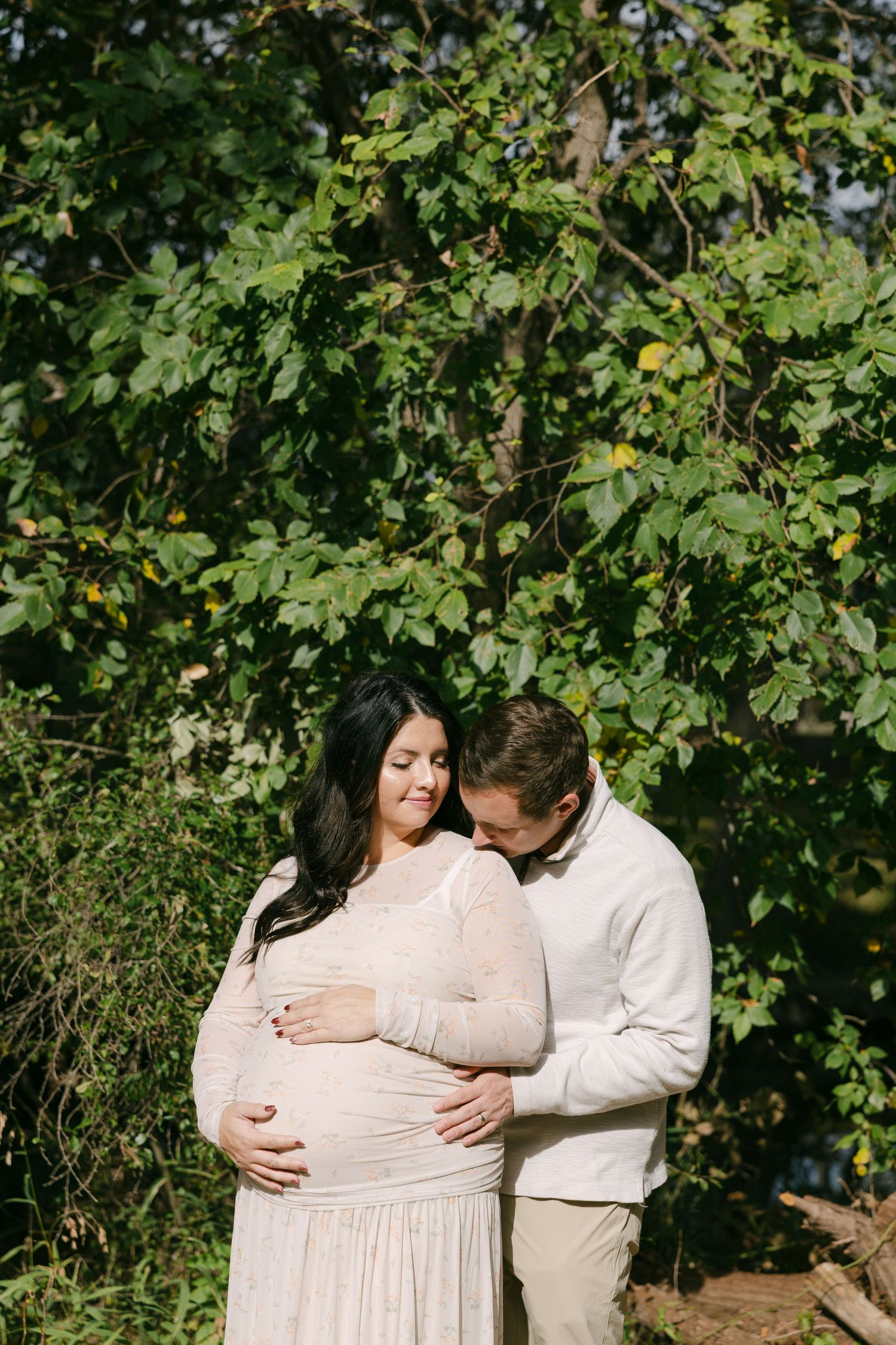 Couple sharing a tender moment outdoors, man kissing pregnant womans head near green trees.