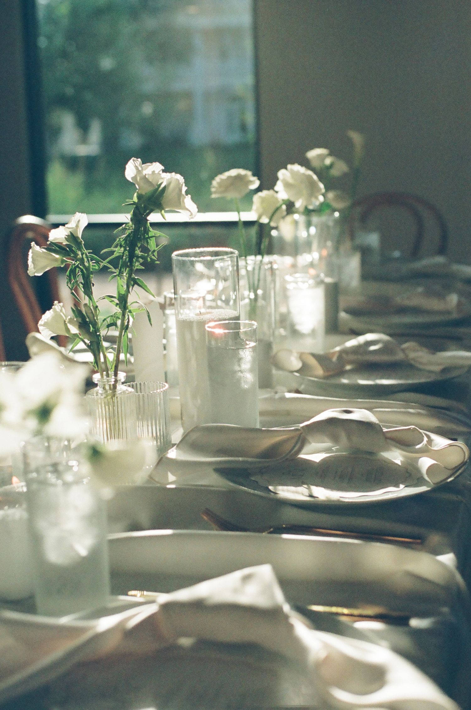 Sunlit wedding table with white ranunculus in bud vases, candles, silver charger plates, and linen napkins by a window.