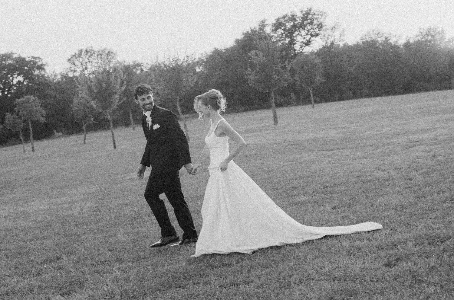 Black and white photo of bride and groom holding hands walking across a green lawn on their wedding day.