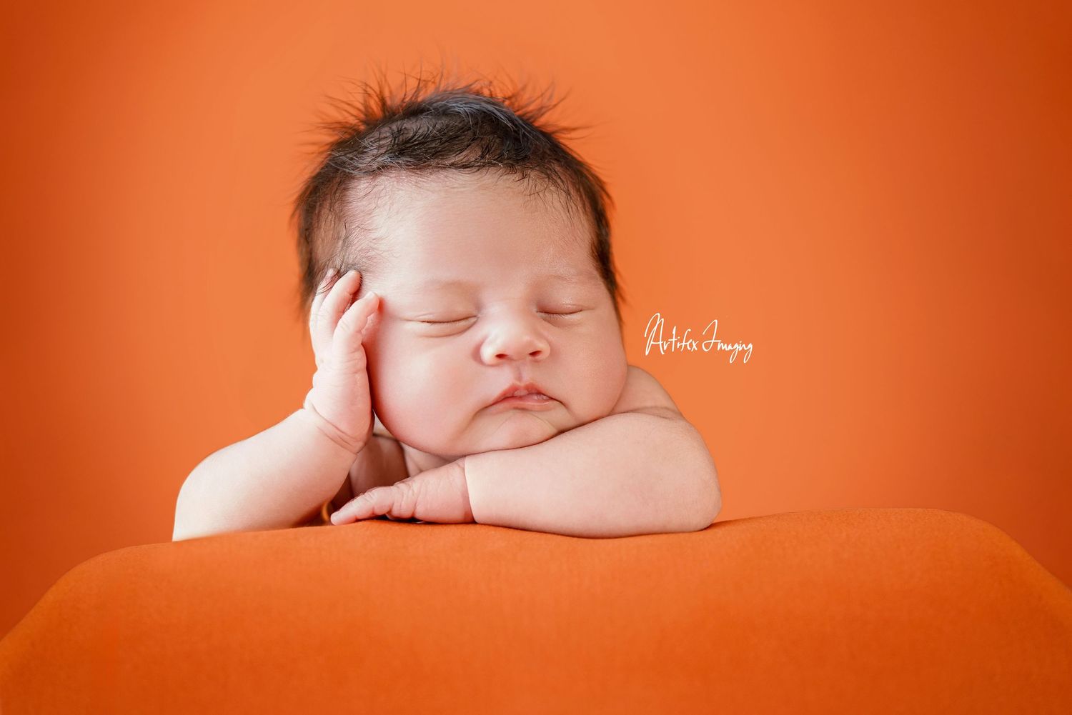 Newborn session featuring a sleeping infant propped up on his elbow in orange fabric setup taken by Artifex Imaging in Bakersfield, CA.