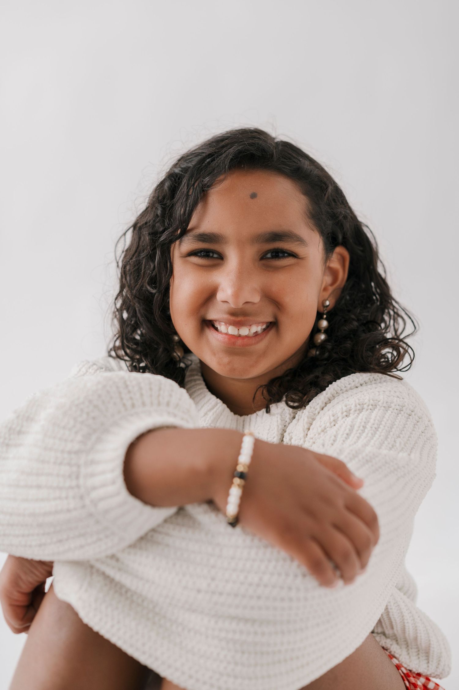 Smiling young woman with curly hair wearing a white knit sweater and beaded bracelet, seated against a white background.