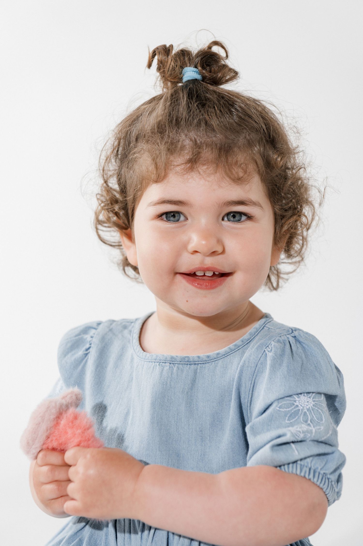 Smiling toddler girl with curly hair in blue dress holding a pink fluffy toy against white background.